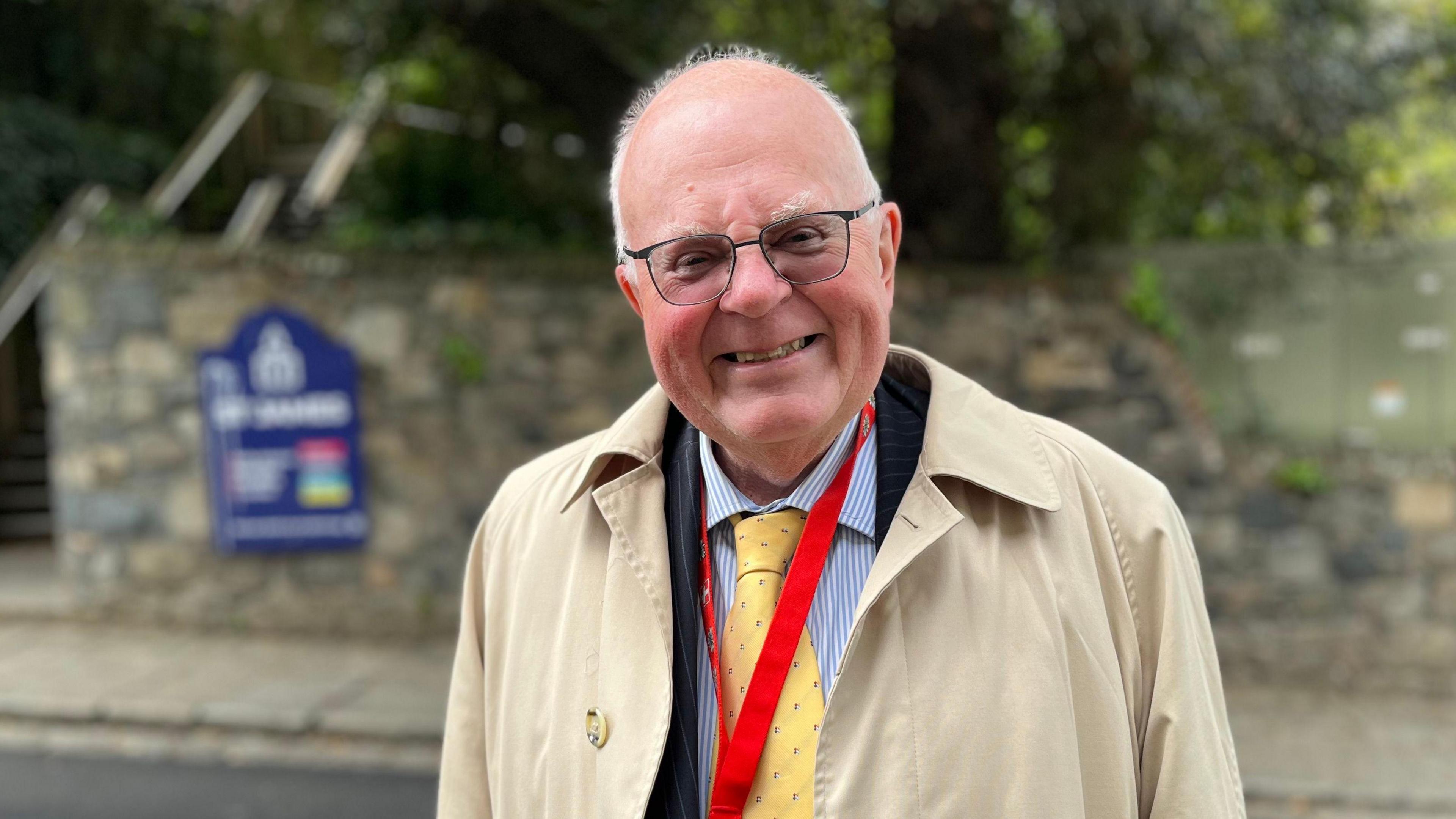 Deputy Chris Le Tissier standing on the side of a road wearing a suit, striped shirt and tie glasses and a beige overcoat smiling at the camera.