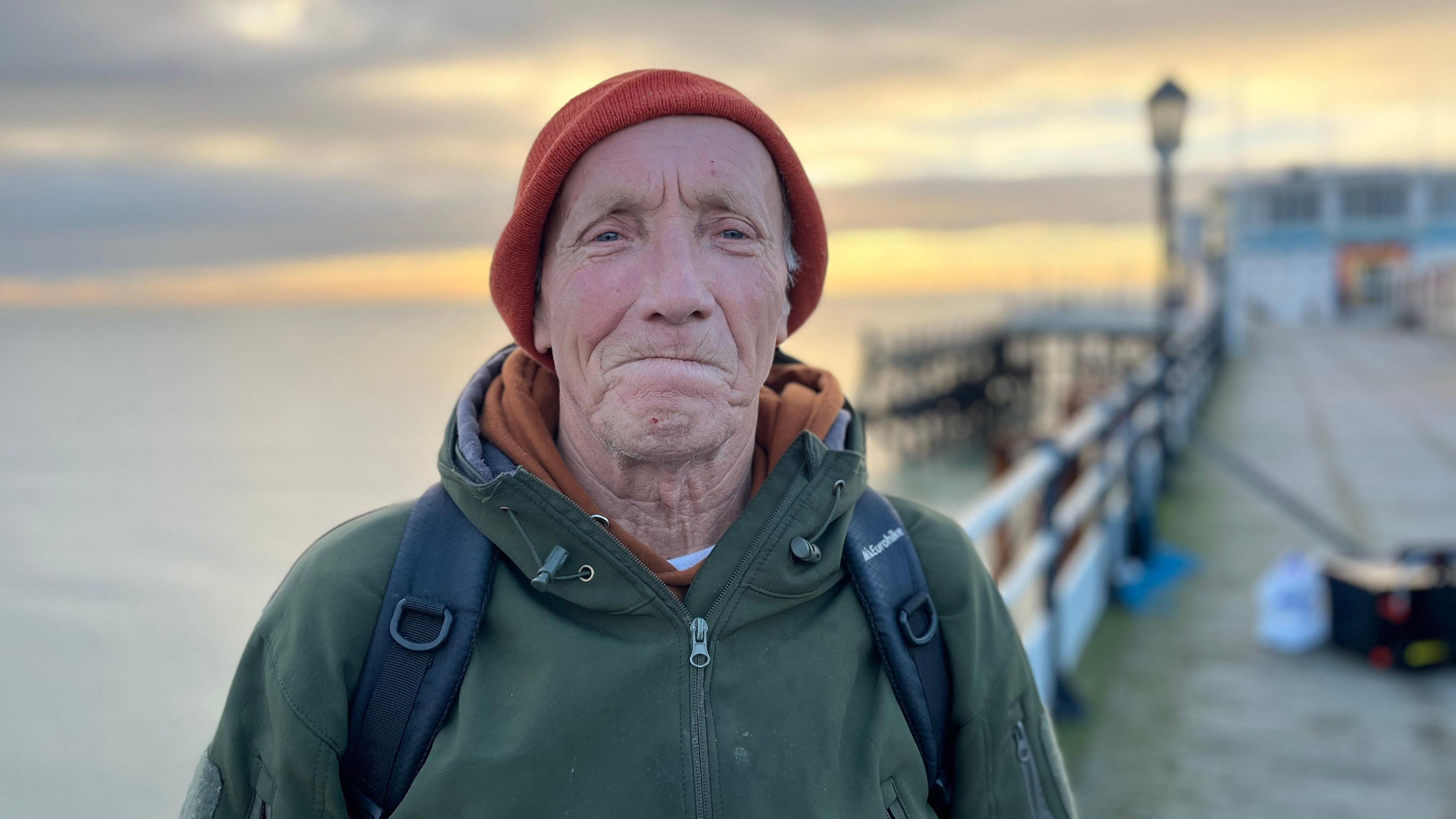 Fisherman Bas standing with his rods behind him smiling at the camera wearing a jacket and beanie 