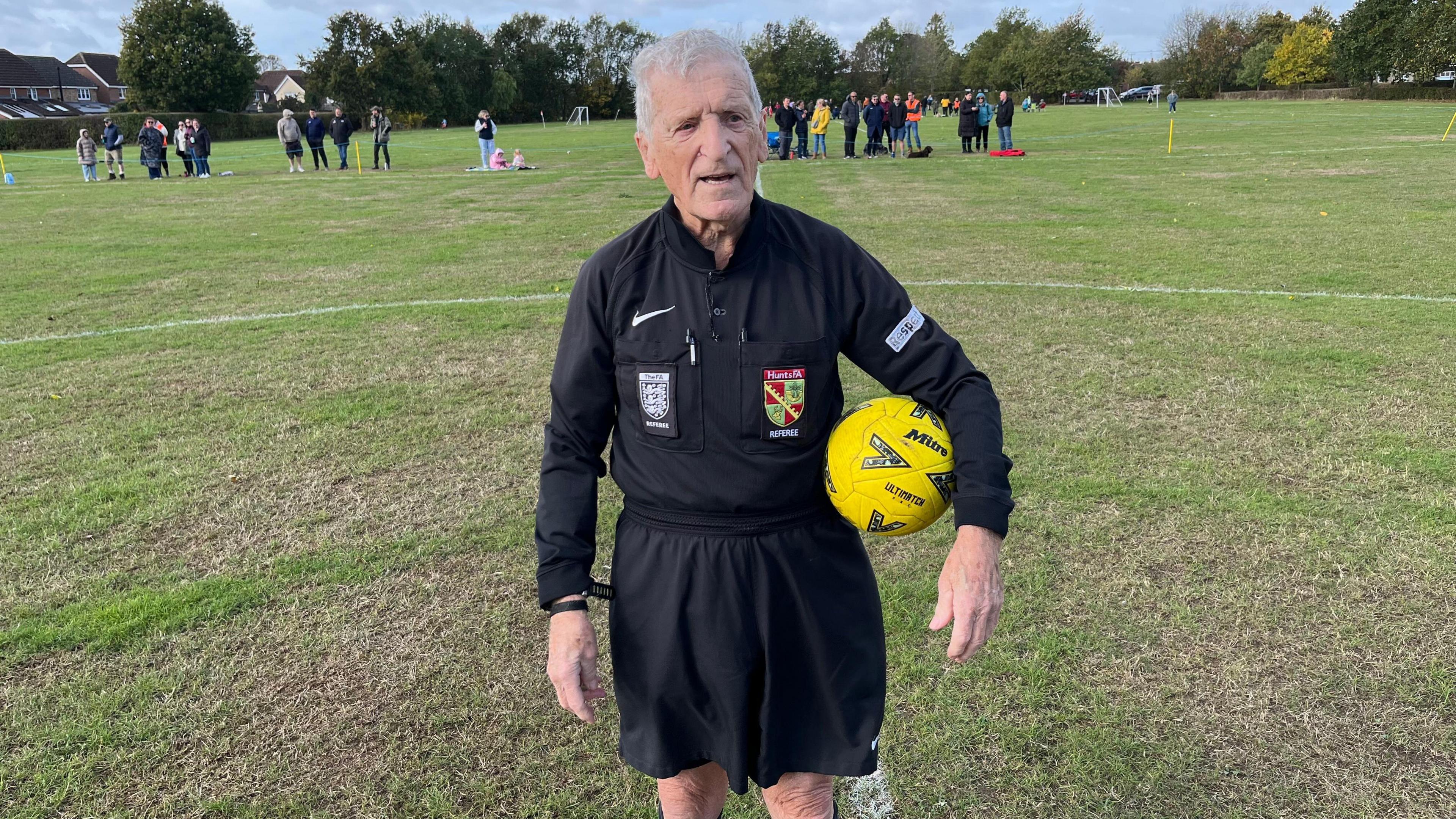 87-year-old football referee Percy Parker stands on a grassy pitch, with a yellow football under his arm. Spectators and players are visible in the background.

