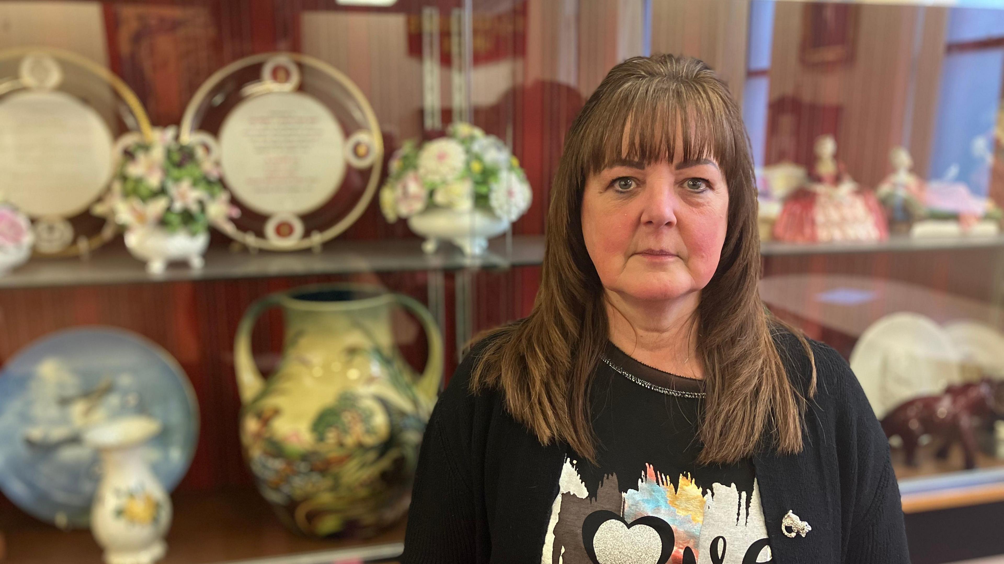 A lady with long brown hair in a black top with a design on and a black cardigan over the top. She is stood in front of various bits of pottery placed on a shelf.