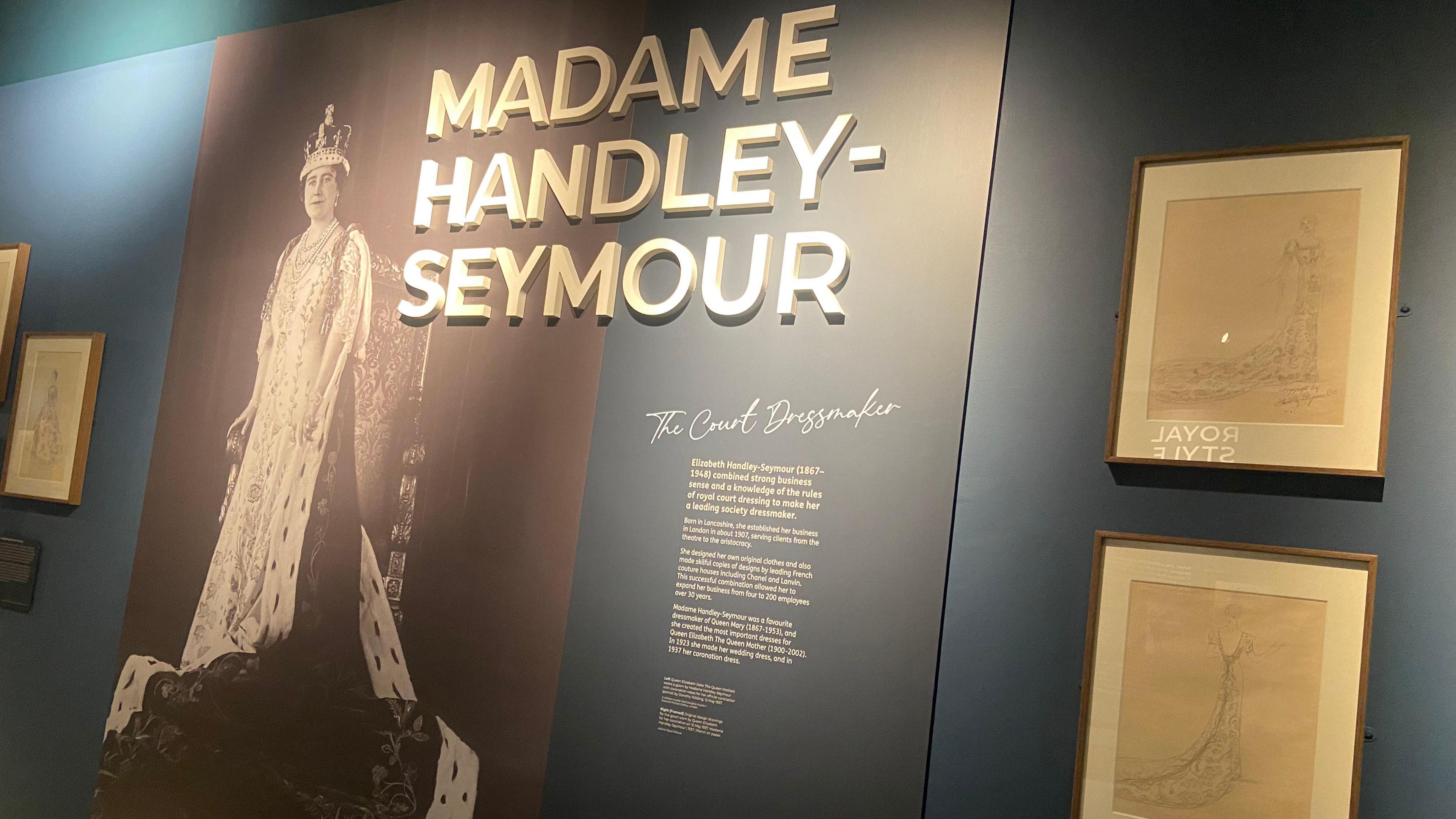 Exhibition sign saying 'Madame Handley-Seymour The Court Dressmaker' A large image of Queen Elizabeth (later The Queen Mother) wearing her coronation gown, robes and crown. Framed sketches are either side.