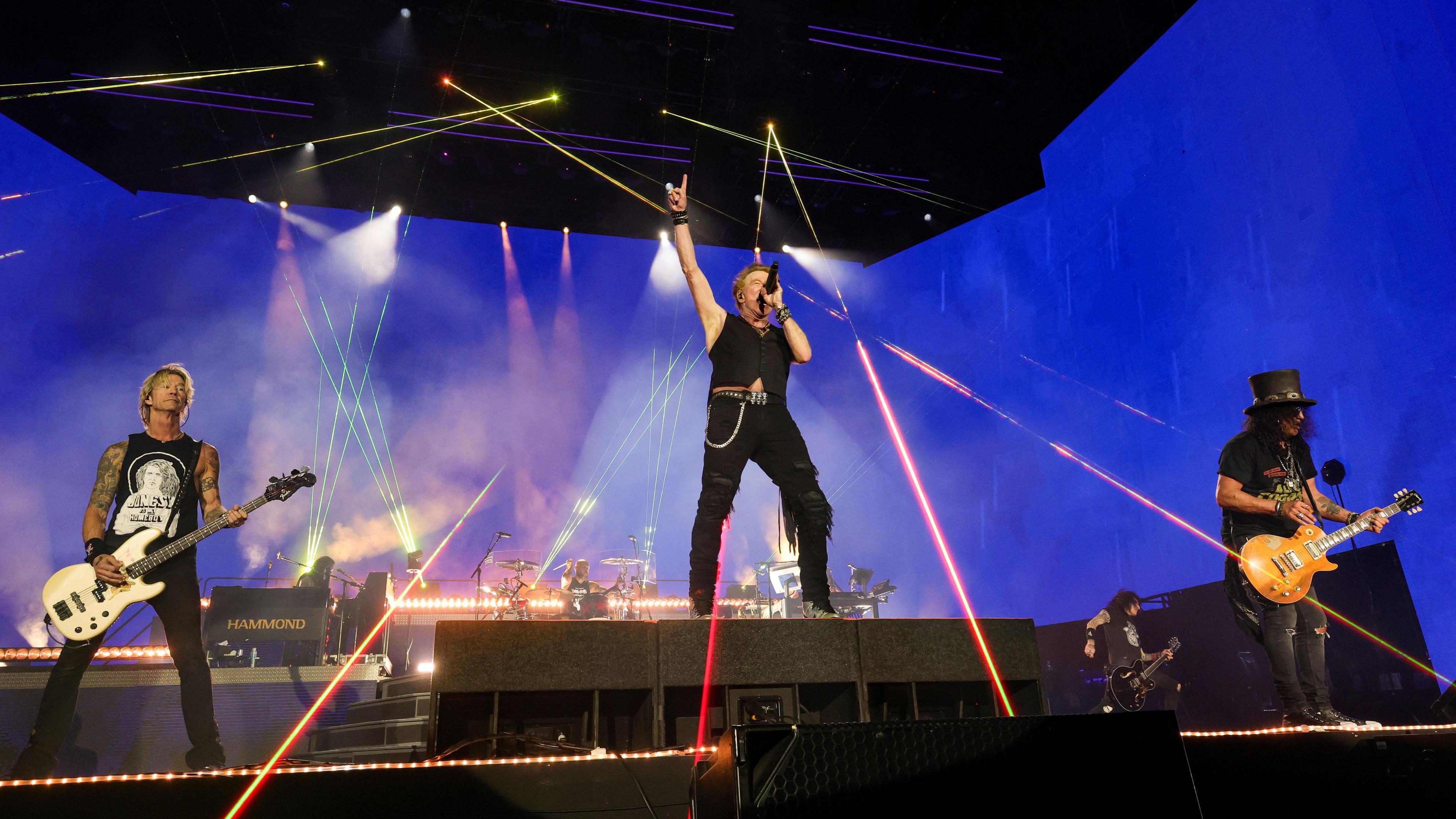 Three members of a rock band on a stage. Red strobe lighting is lighting up the stage. The lead singer, a man in black trousers and a black waistcoat is jumping up in the air and two guitarists are standing on either side of him.