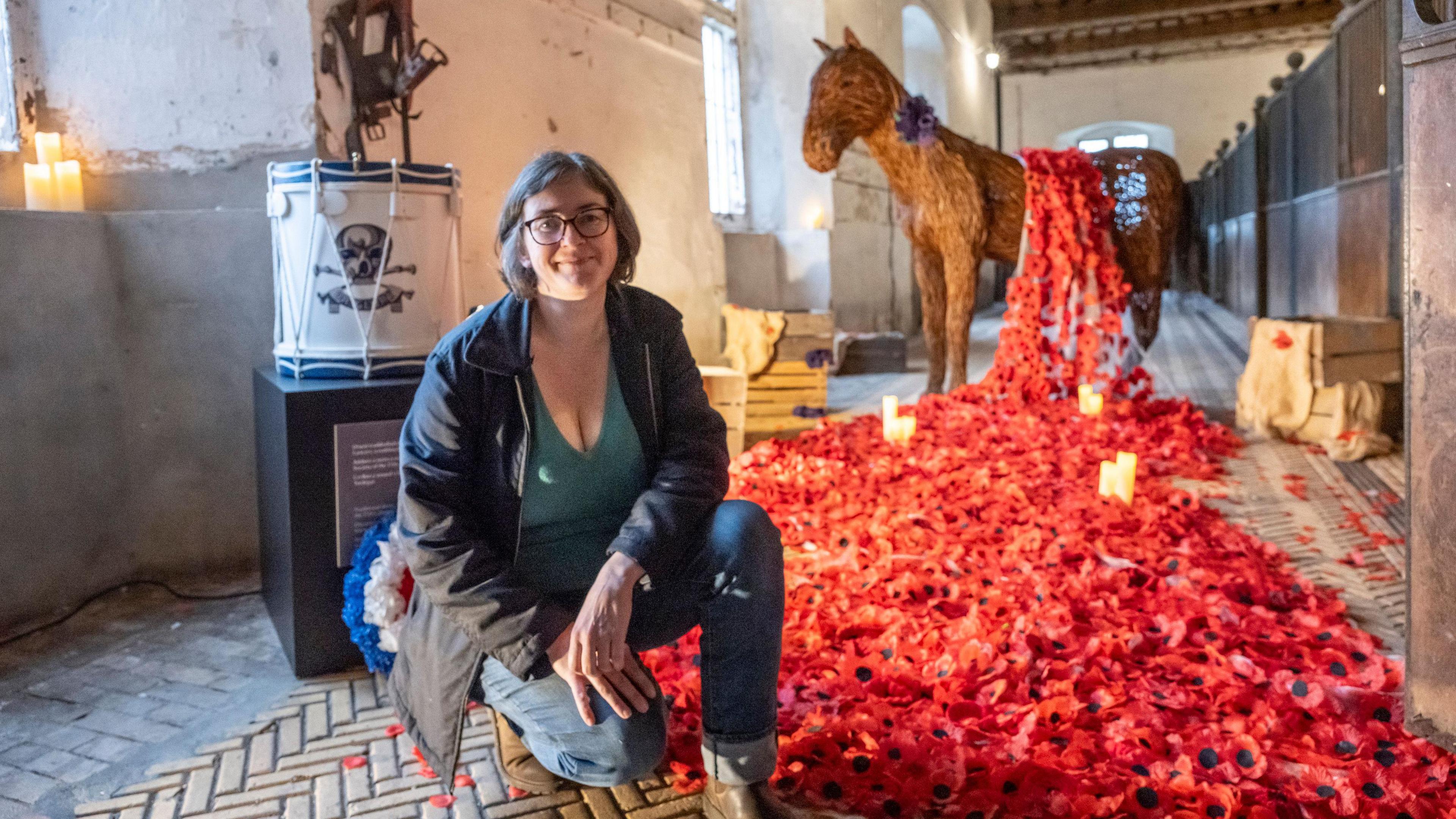 An exact replica of Godfrey Morgan’s military horse ‘Sir Briggs’, which has been hand-woven by local willow artist Sarah Hatton, is in the background with a trail of more than four thousand poppies leading up to and then wrapped around it. Sarah Hatton rest on one knee on the foreground smiling at the camera 