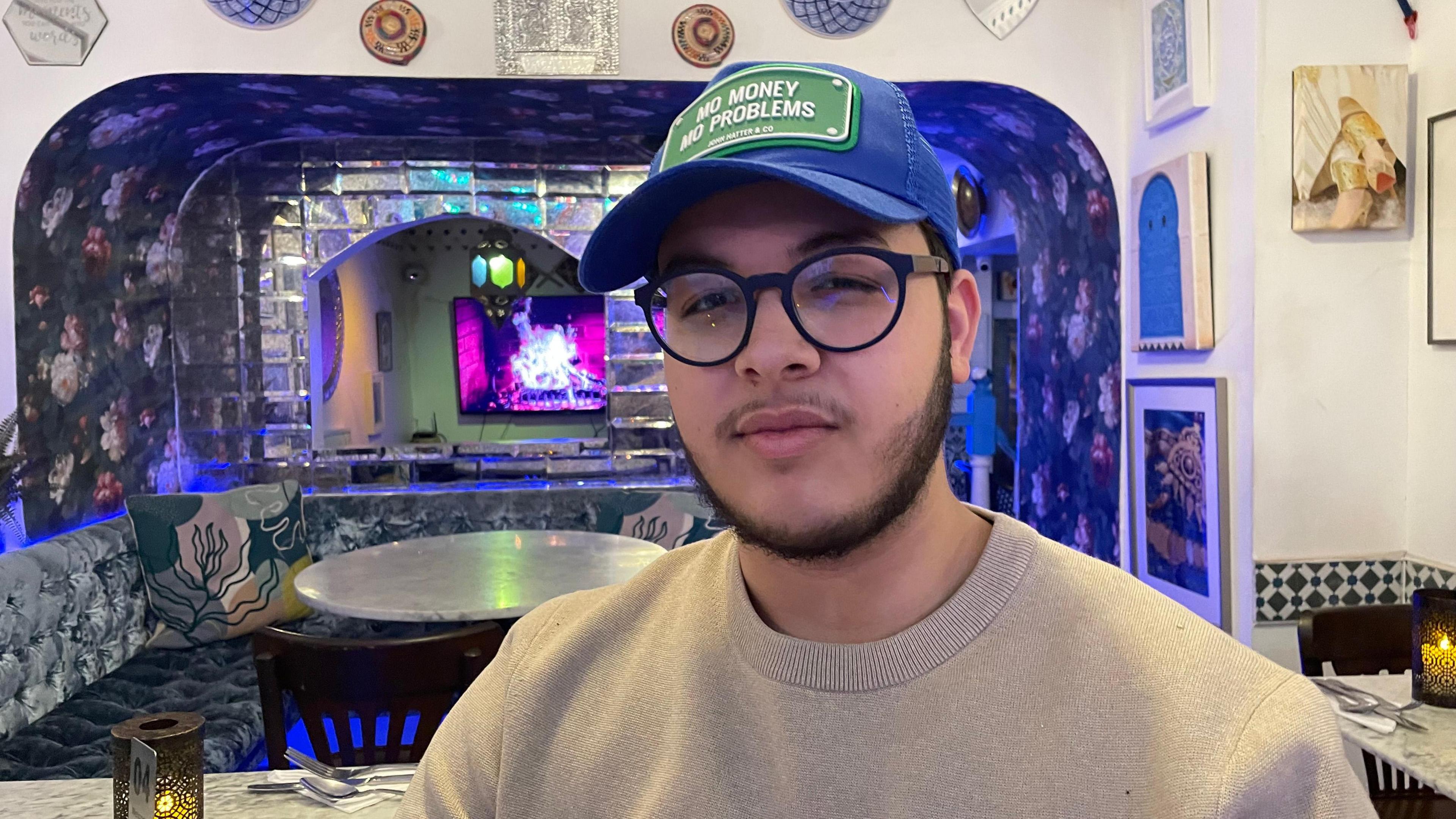 Burrë me syze dhe mjekër me një kapelë blu dhe jeshile që thotë "pa para, pa probleme" me shkronja të bardha