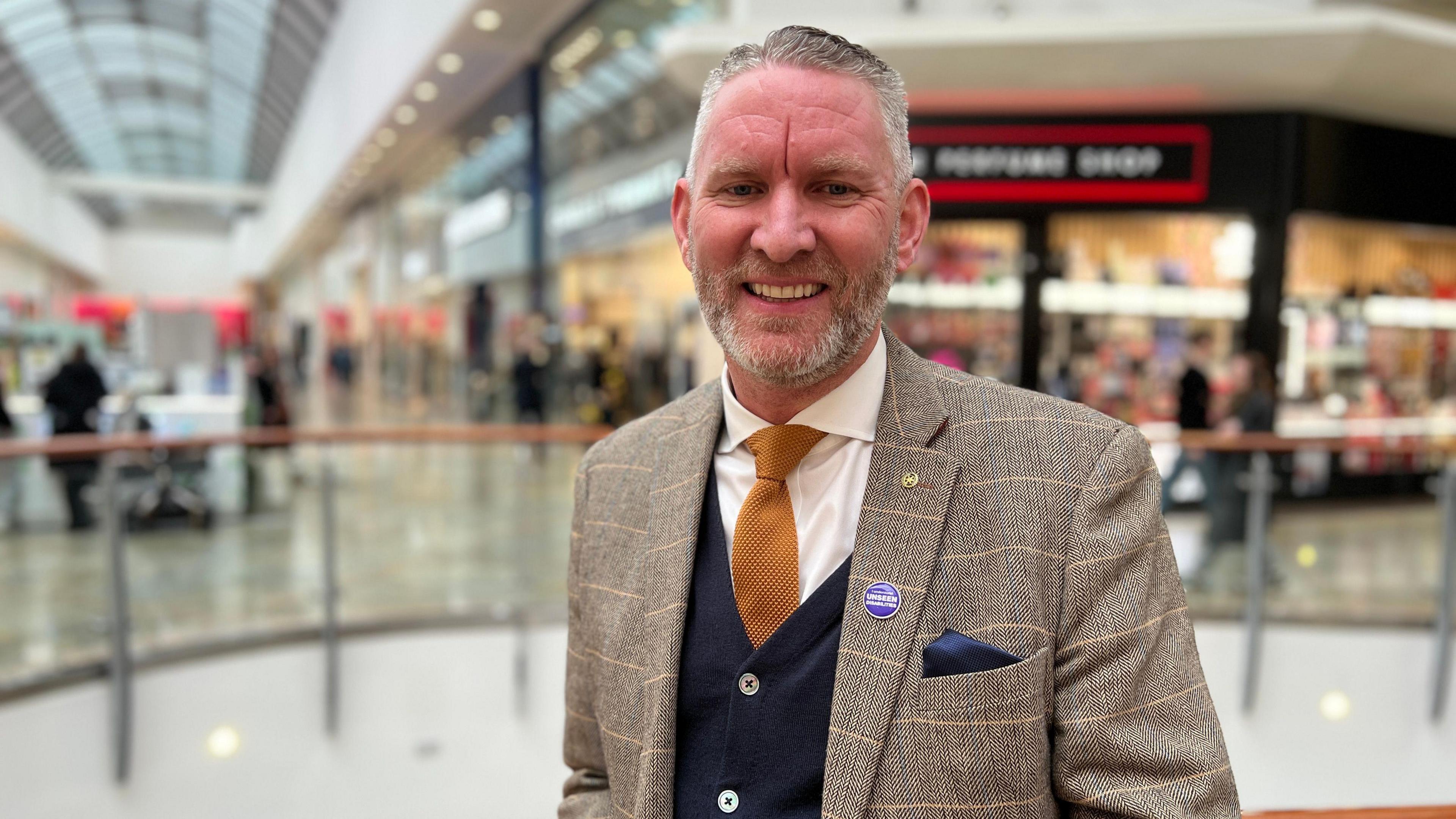 A head-and-shoulders photo of a man with short grey hair and grey stubble, wearing a brown blazer, a yellow tie and a blue cardigan, smiling at the camera. He's standing inside a shopping centre with shops behind him.