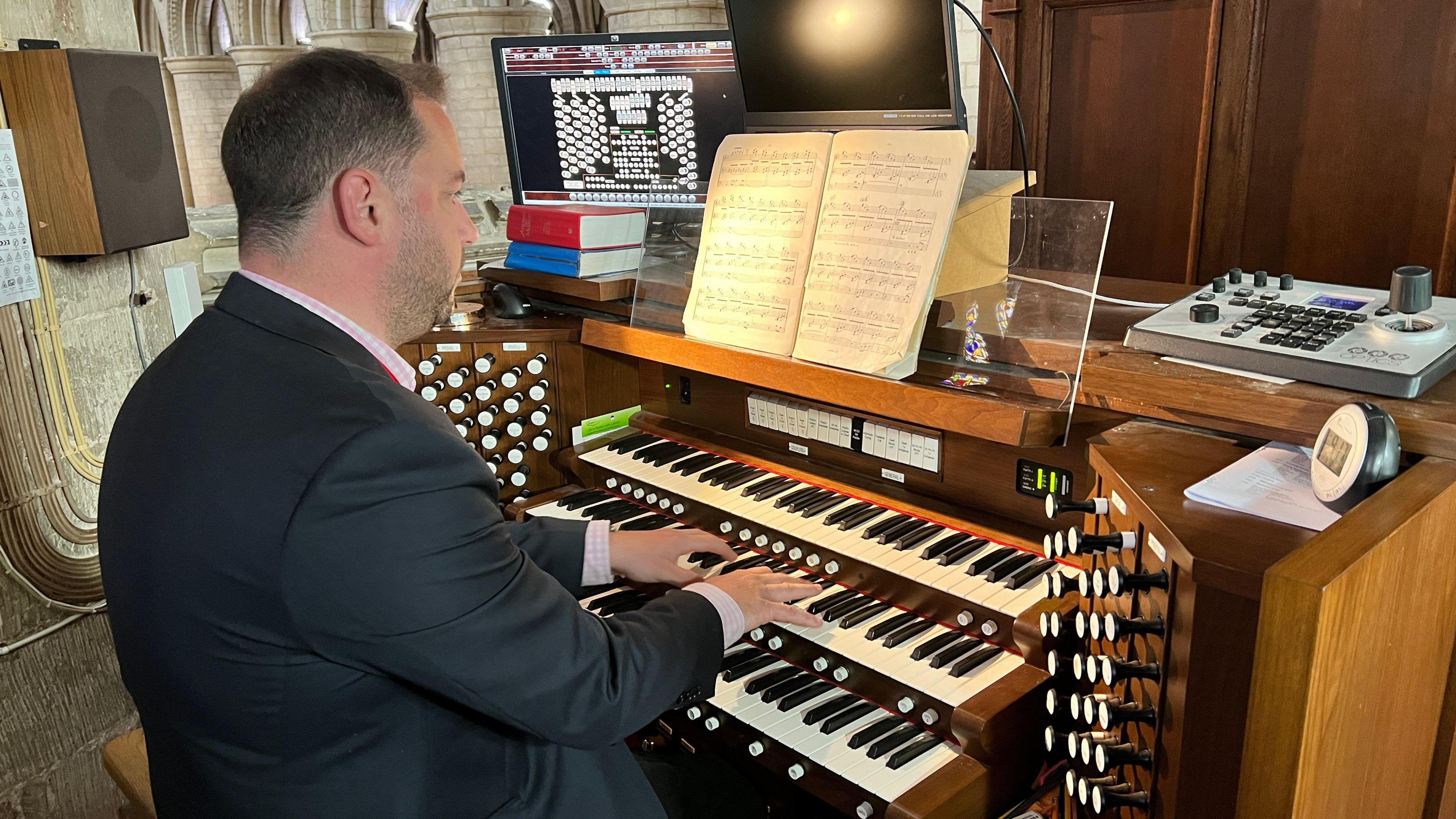 A man in a suit plays a large pipe organ console inside a cathedral. The console includes multiple keyboards, numerous stops, and various controls. Open sheet music rests on the stand, flanked by two computer monitors displaying digital scores. Behind him, the cathedral’s ornate stone architecture and arched columns are visible.