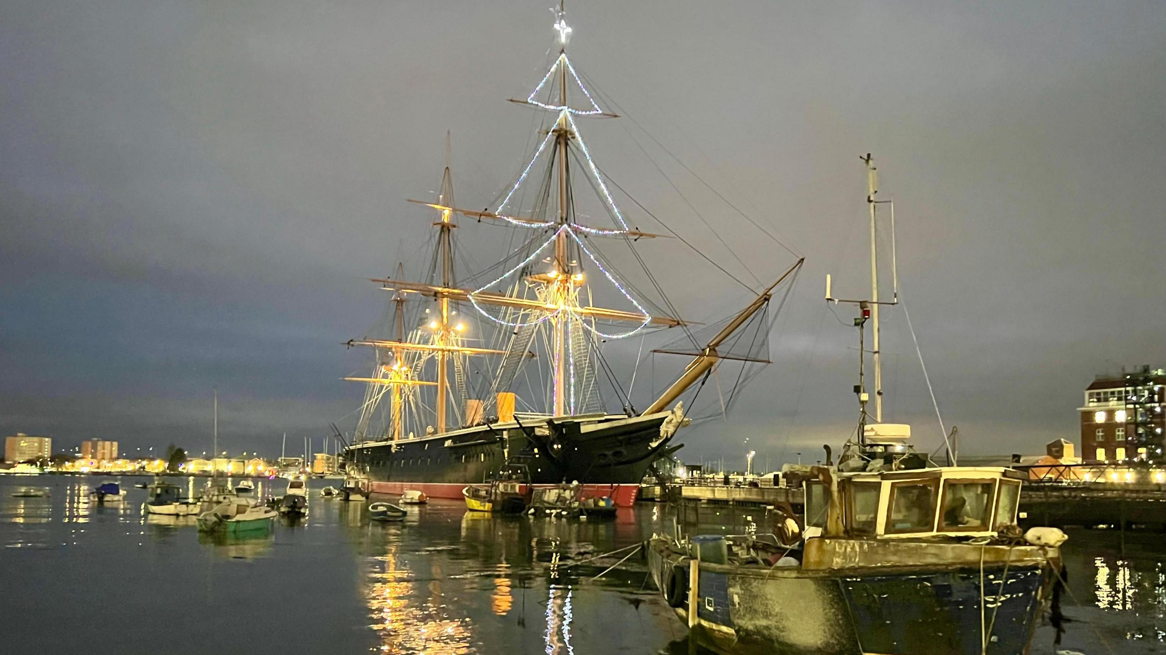 HMS Warrior is a warship built for the Royal Navy in the 19th Century. It has a black and red hull. The front mast has Christmas lights in shaped like a Christmas tree. The sun has set and lights can be seen illumination boats and buildings around the harbour.