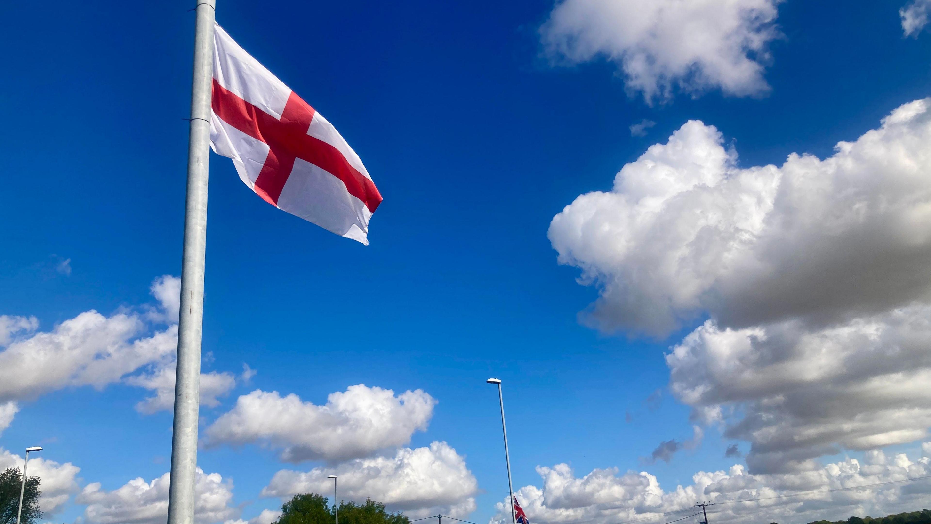 A St George's flag hangs from a lamppost against a blue sky with white clouds.
