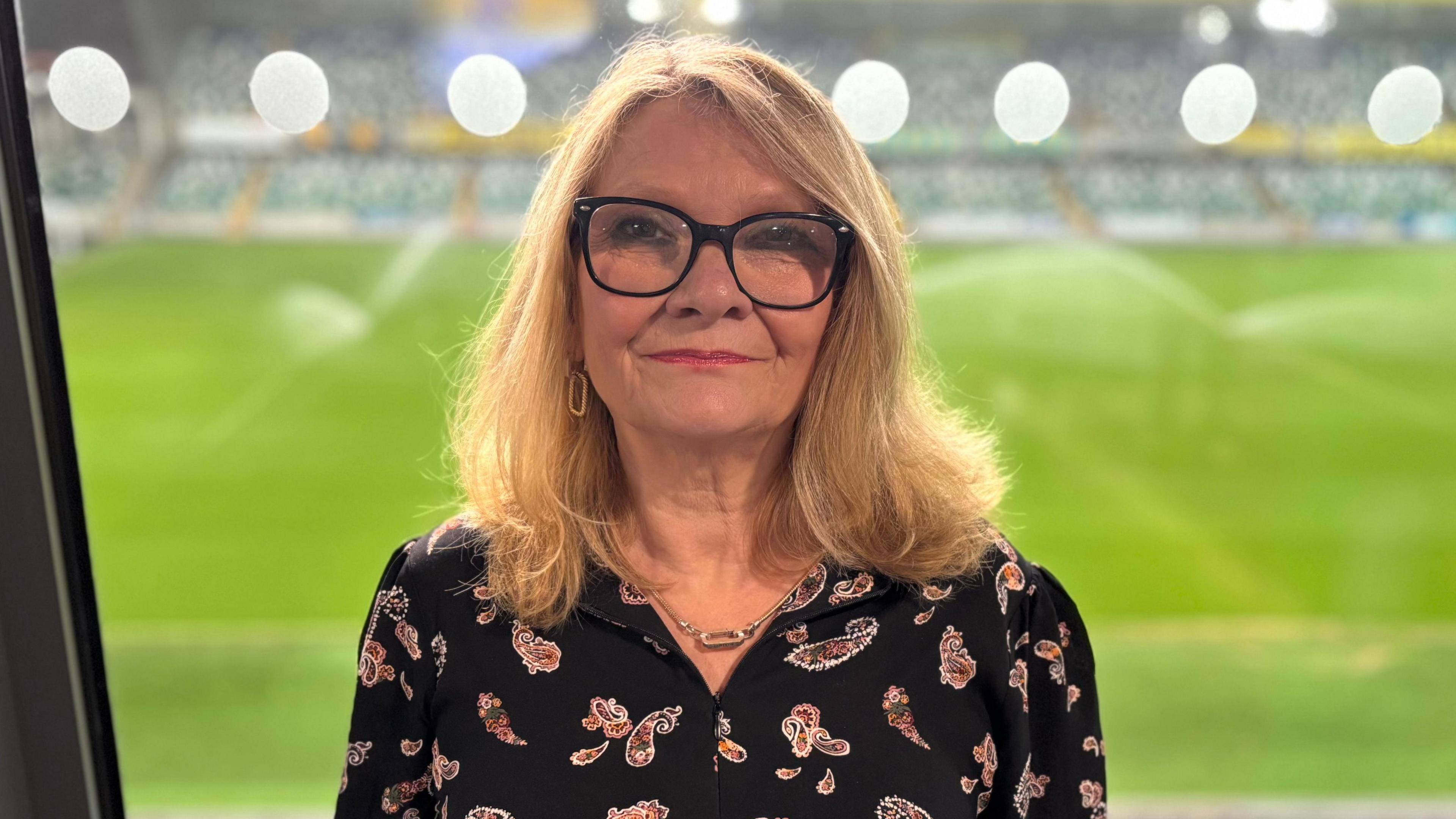Sarah Mason is standing in a glass room overlooking a green football pitch. She has long blonde hair, is wearing black framed glasses and a black paisley red patterened top. 