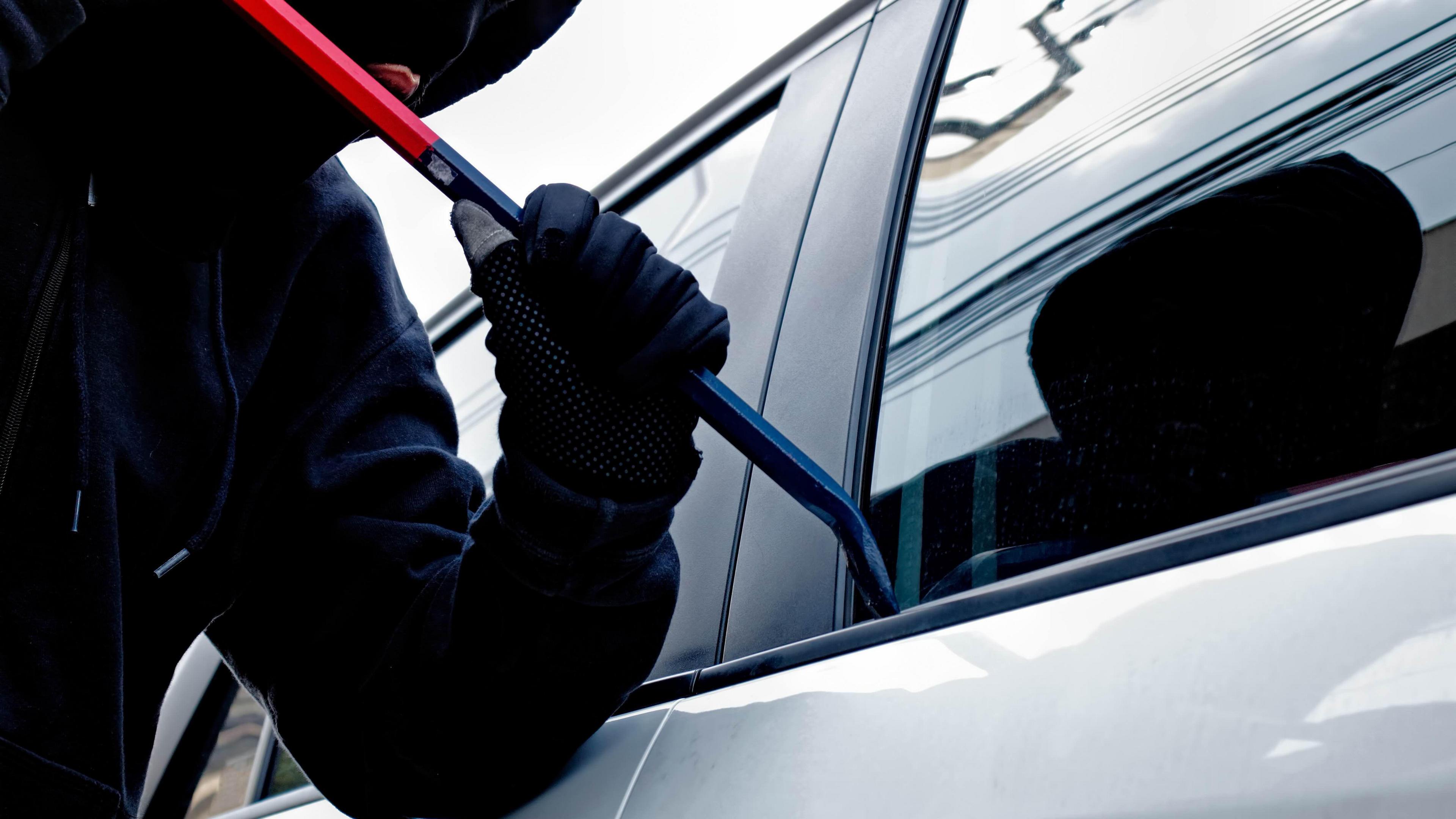 A masked person wearing black trying to break into a car with a long  blue and red metal stick