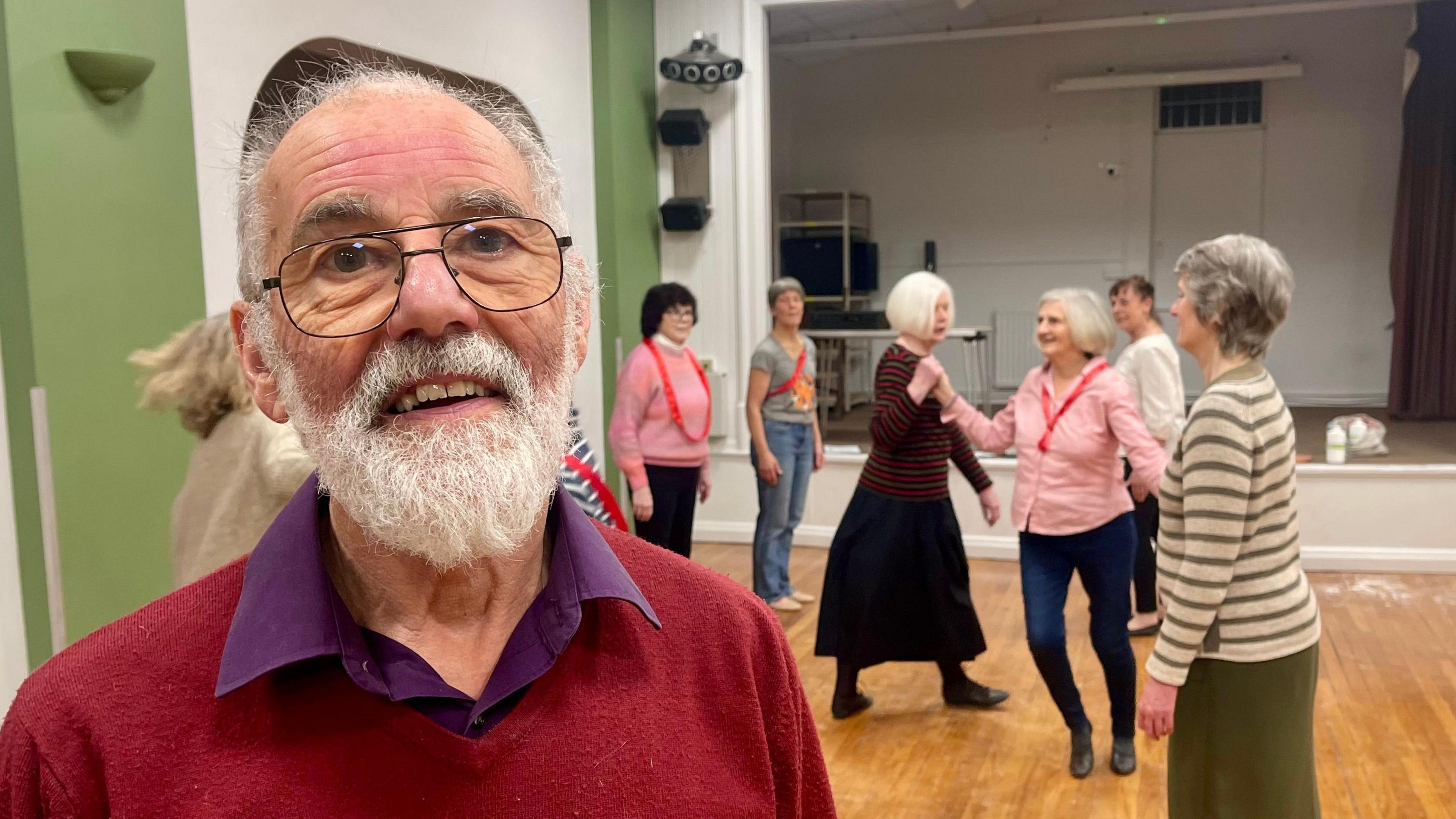 A man with a white beard, balding head, glasses, maroon jumper and purple shirt is standing in a village hall. Behind him are six female dancers, some wearing red sashes. Two are holding hands in a traditional dance move.