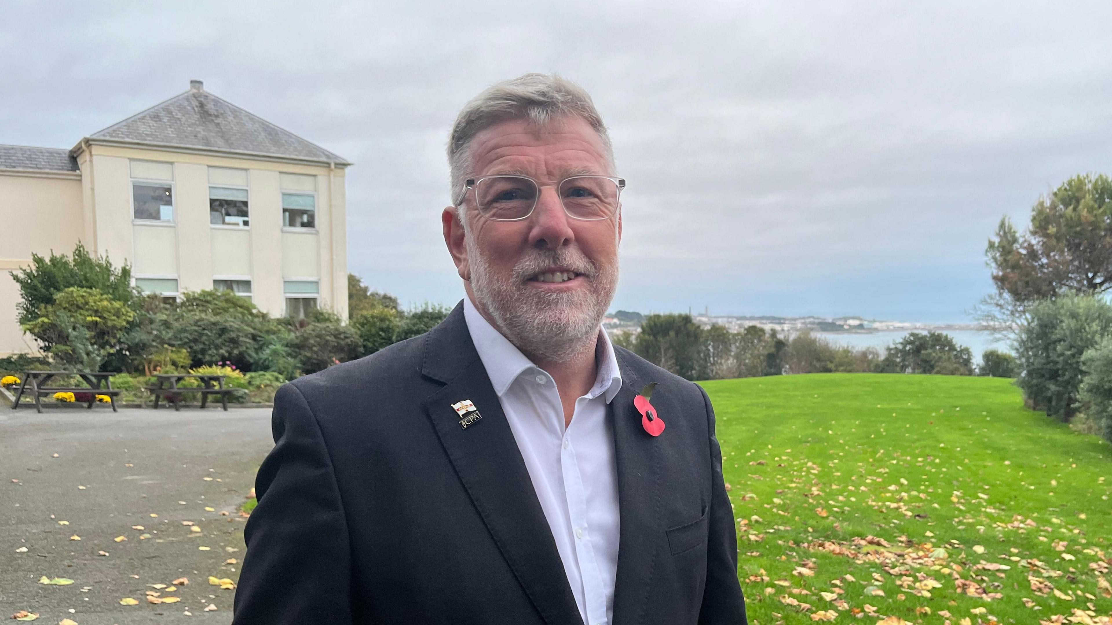 Deputy Adrian Gabriel wearing a black suit blazer and white shirt. He has short grey hair and a short grey beard. He is wearing glasses. He is stood in the grounds of a building looking over the Guernsey coast. 