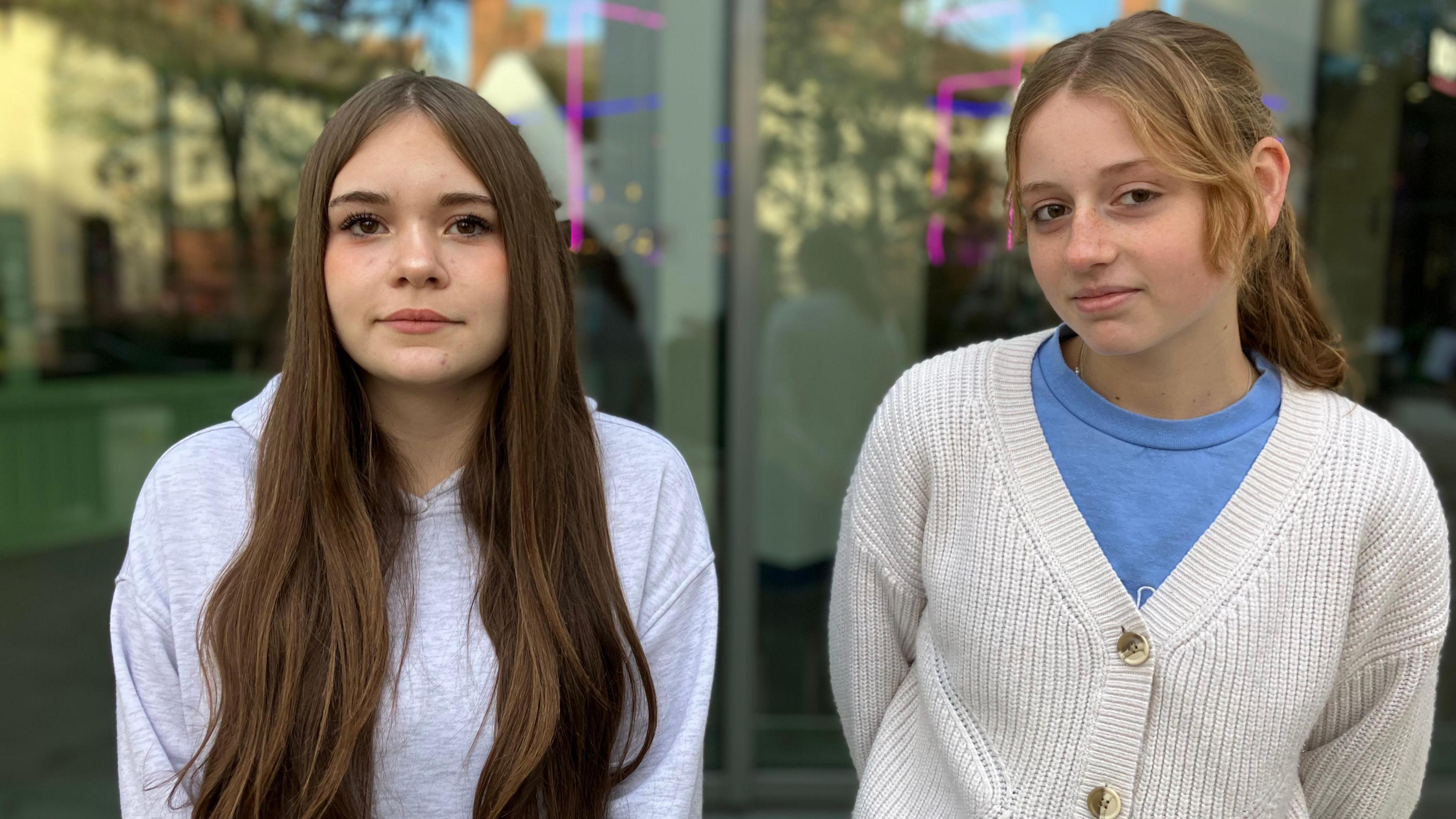 Two teenage girls stand in front of a glass shop window.