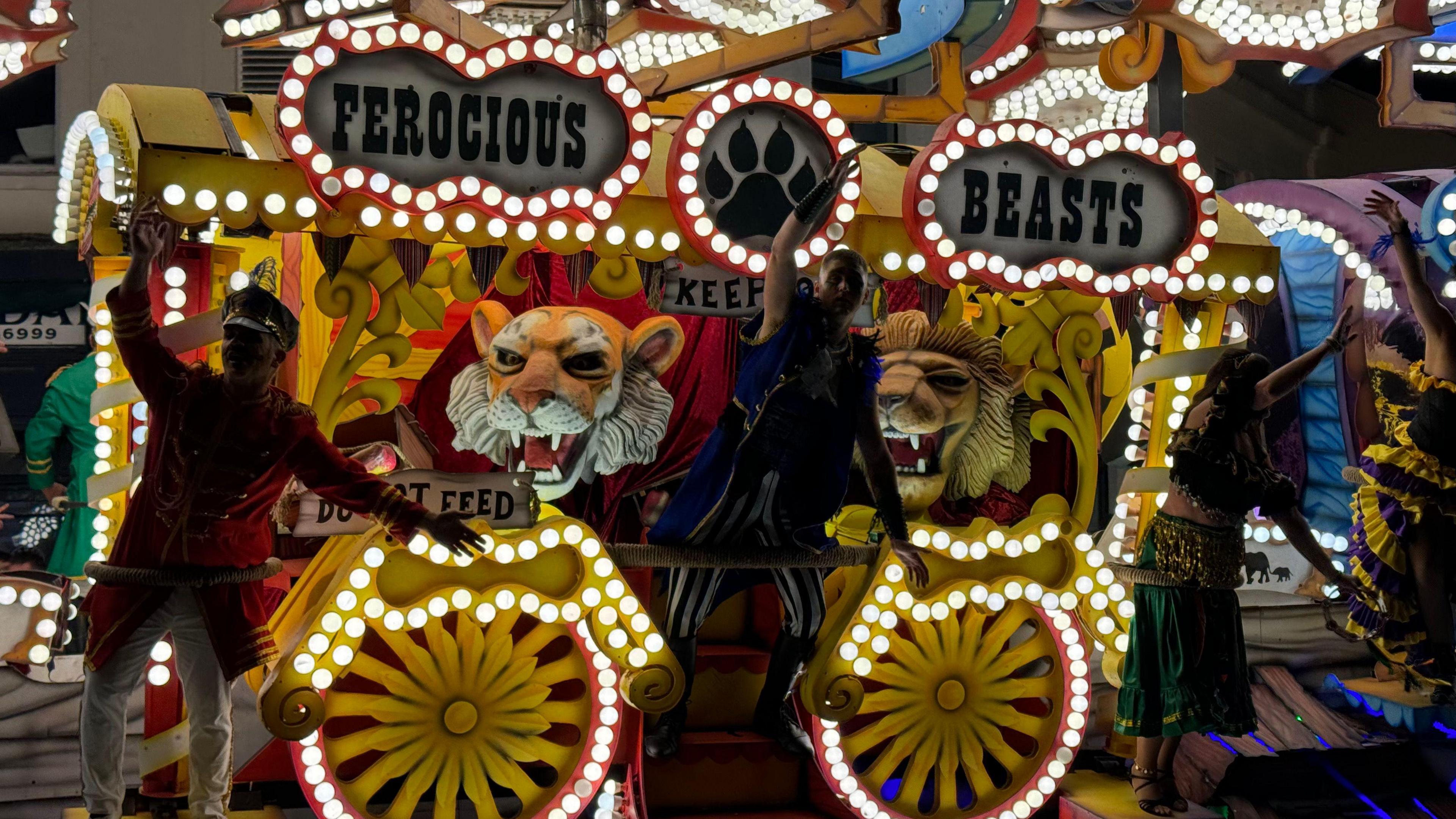 A carnival float which says 'ferocious beasts'. The cart features two lion heads and three people stand on the edge of the cart waving at the crowd