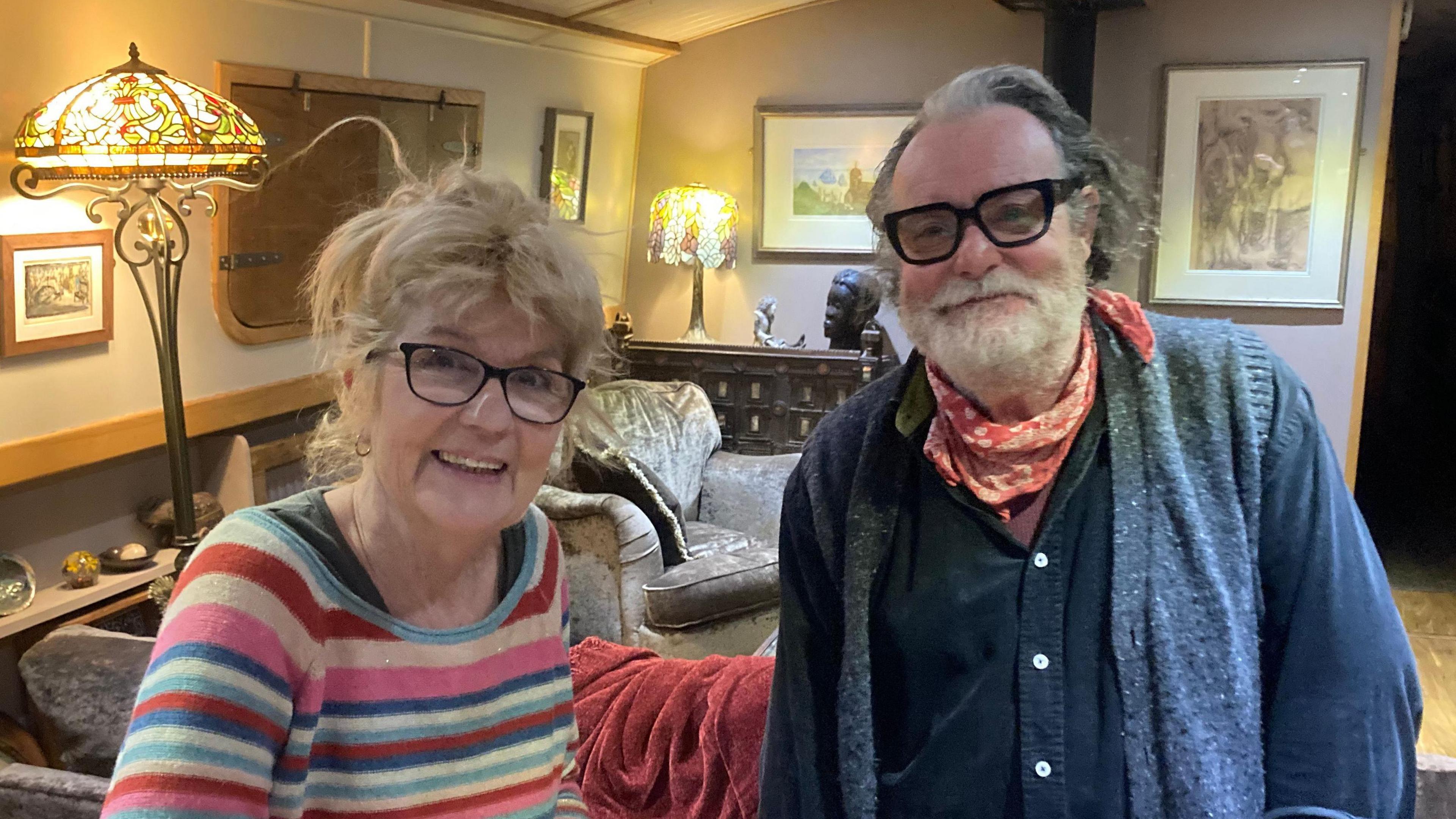 A woman wearing a colourful stripy jumper and her husband, in a blue cardigan, stand inside a living room. There are two lamps behind them, one closer to the lens, with a multi-coloured glass lampshade and pictures on the wall with an armchair visible behind them.