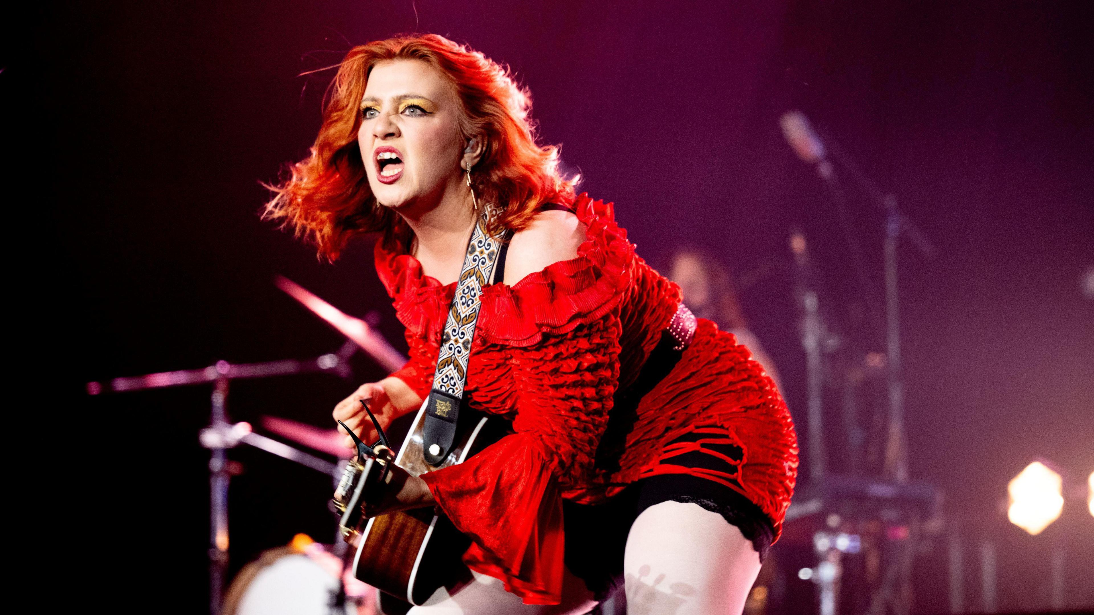 CMAT, a woman with auburn hair plays a guitar on stage. She is wearing a red dress. Behind her out of focus, a drummer can be seen playing.