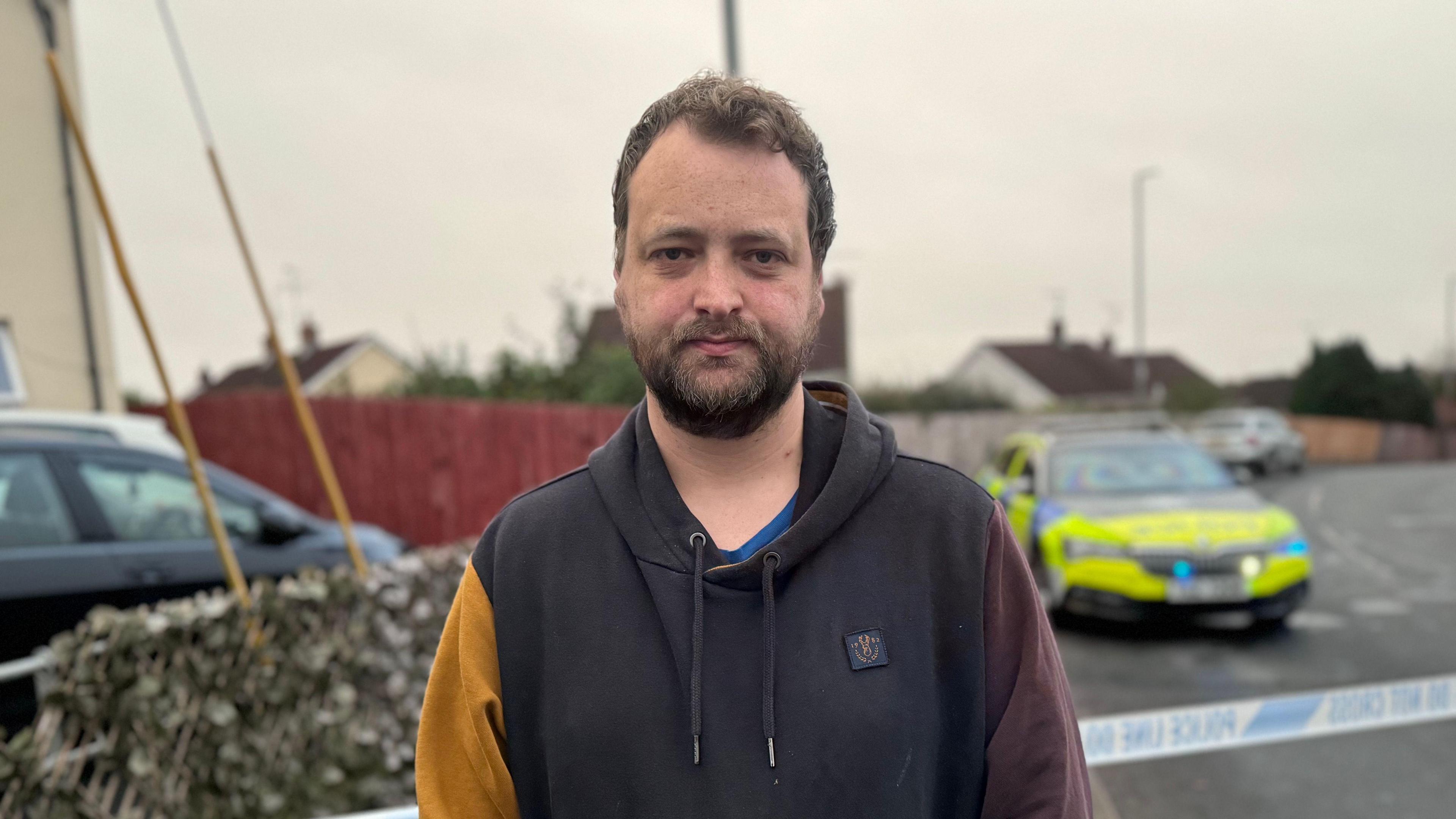 A man with brown hair looks at the camera. He is wearing a hoodie which has a black body, a burnt orange right sleeve, and a dark purple left sleeve. In the background, there is police tape, as well as a PSNI car.