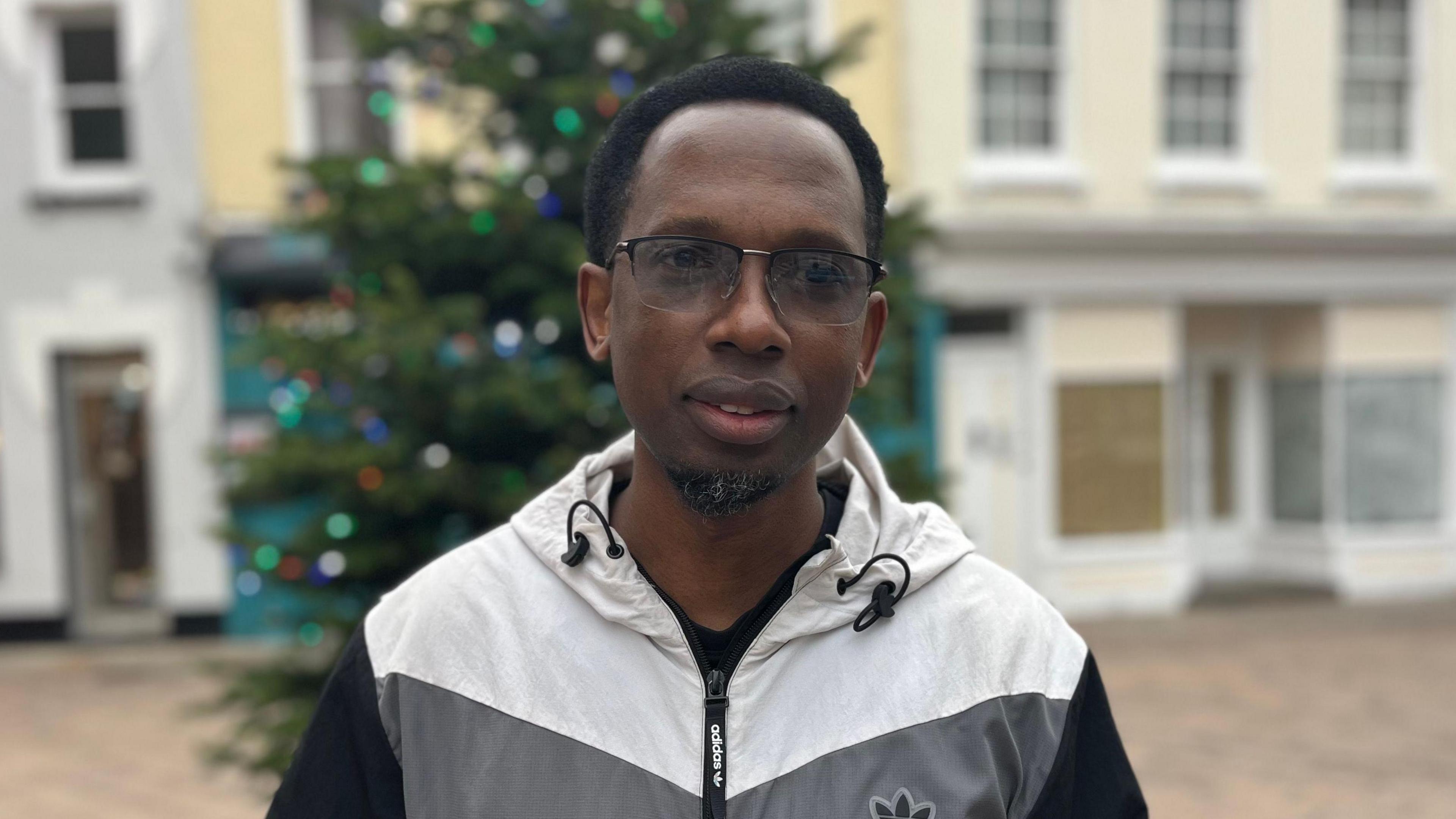 Njabulo Sibanda is a man wearing glasses and a grey, black and white waterproof. He is staring at the camera in front of an out-of-focus Christmas tree and some shops.