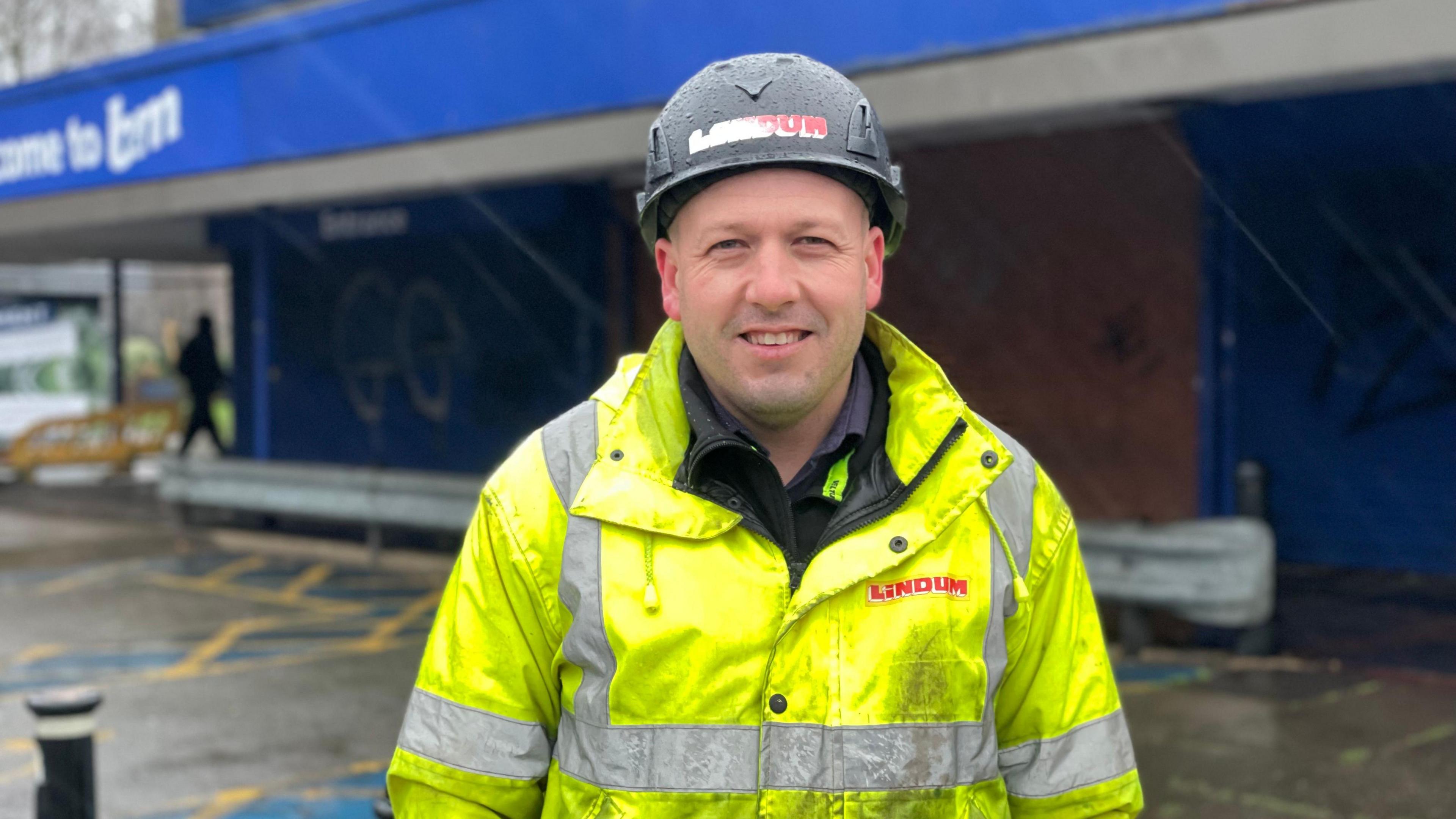 A photo of Lindum Constructions site manager Conrad Cousins. He is wearing a Lindum branded yellow hi-vis jacket and a black hard hat. He is stood in front of the B&M store that is due to be demolished. 