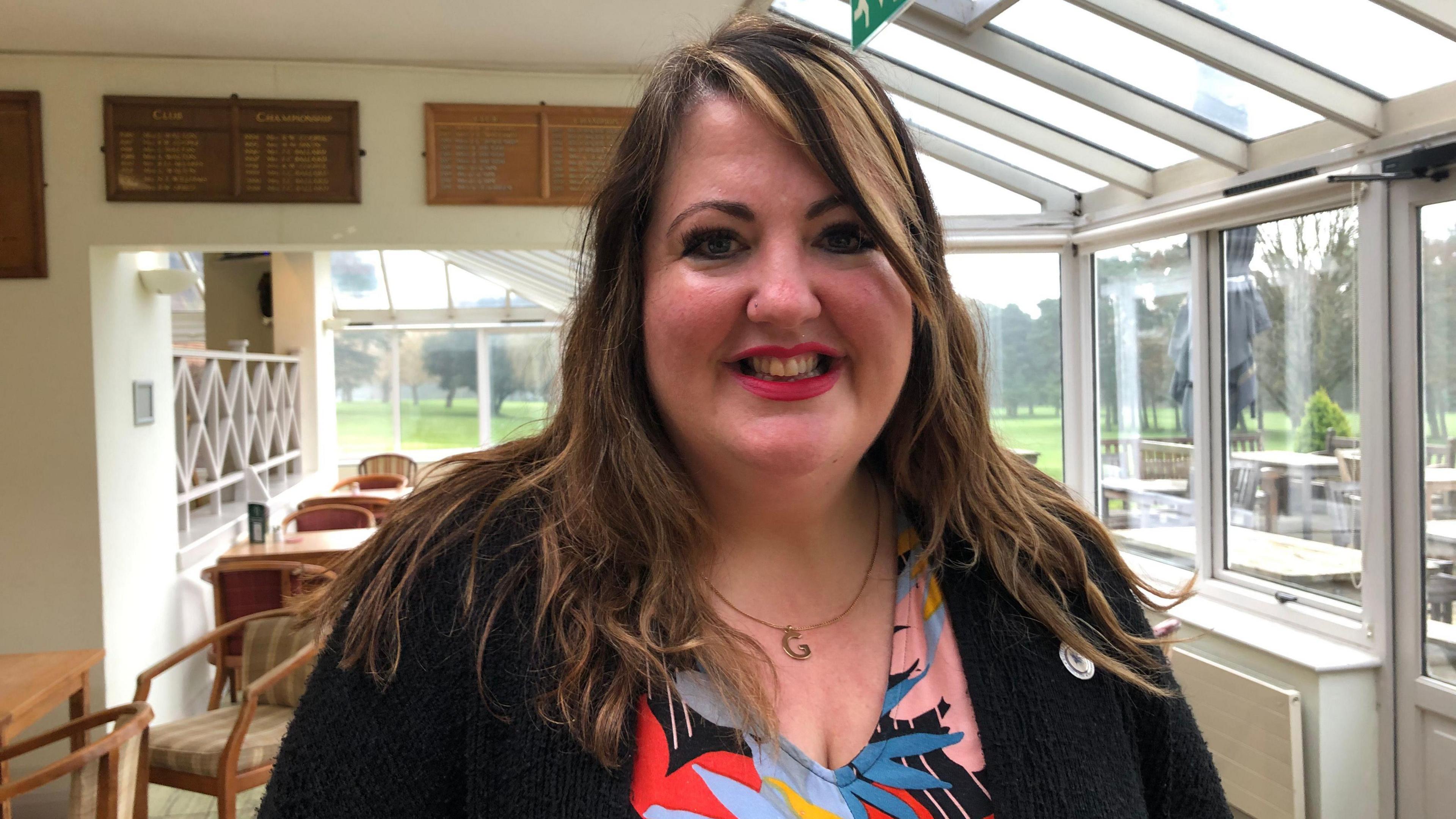 Gini Hackett is in the centre of the frame. She is smiling and is wearing bright lipstick.  She has long dark hair over her shoulders with lighter highlights. She is wearing a bright floral dress and black cardigan.  She is standing in the conservatory area of a restaurant with chairs and tables behind.