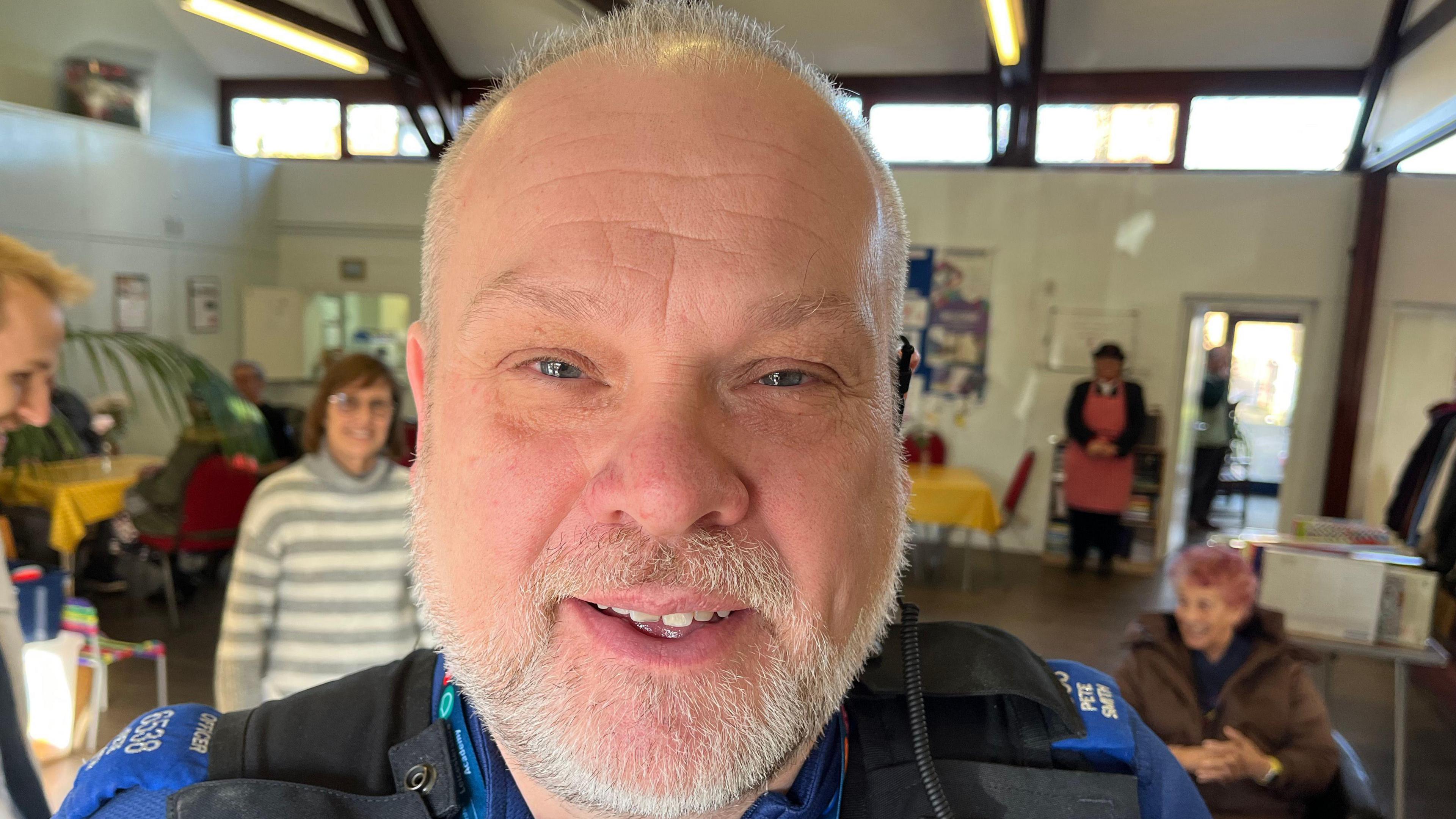 Pete wears a PCSO uniform and smiles at the camera with people in the community centre behind him. 