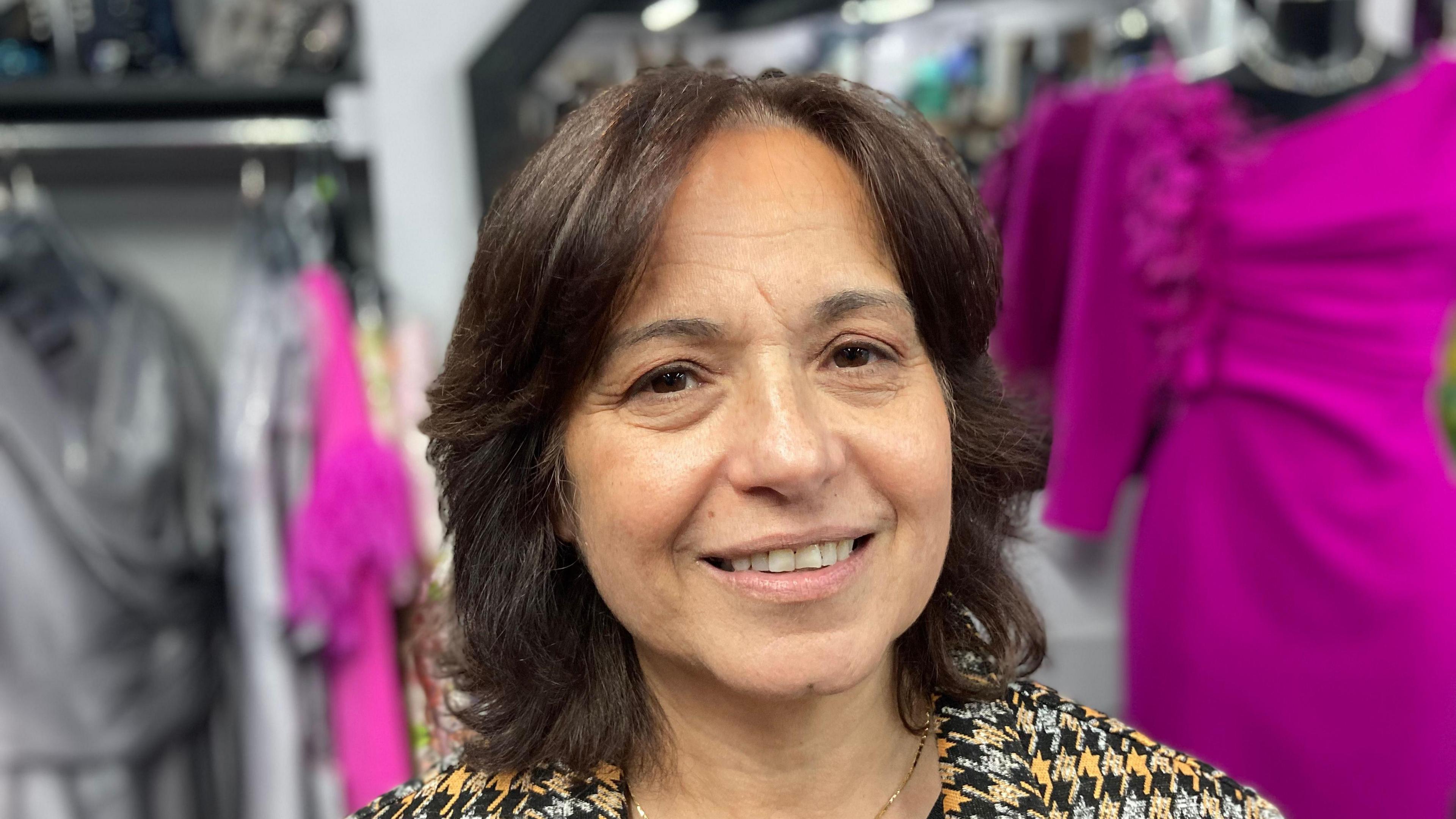 A woman in a black, silver, and yellow top stands in a shop smiling at the camera. There are clothes hanging up behind her, including a purple dress