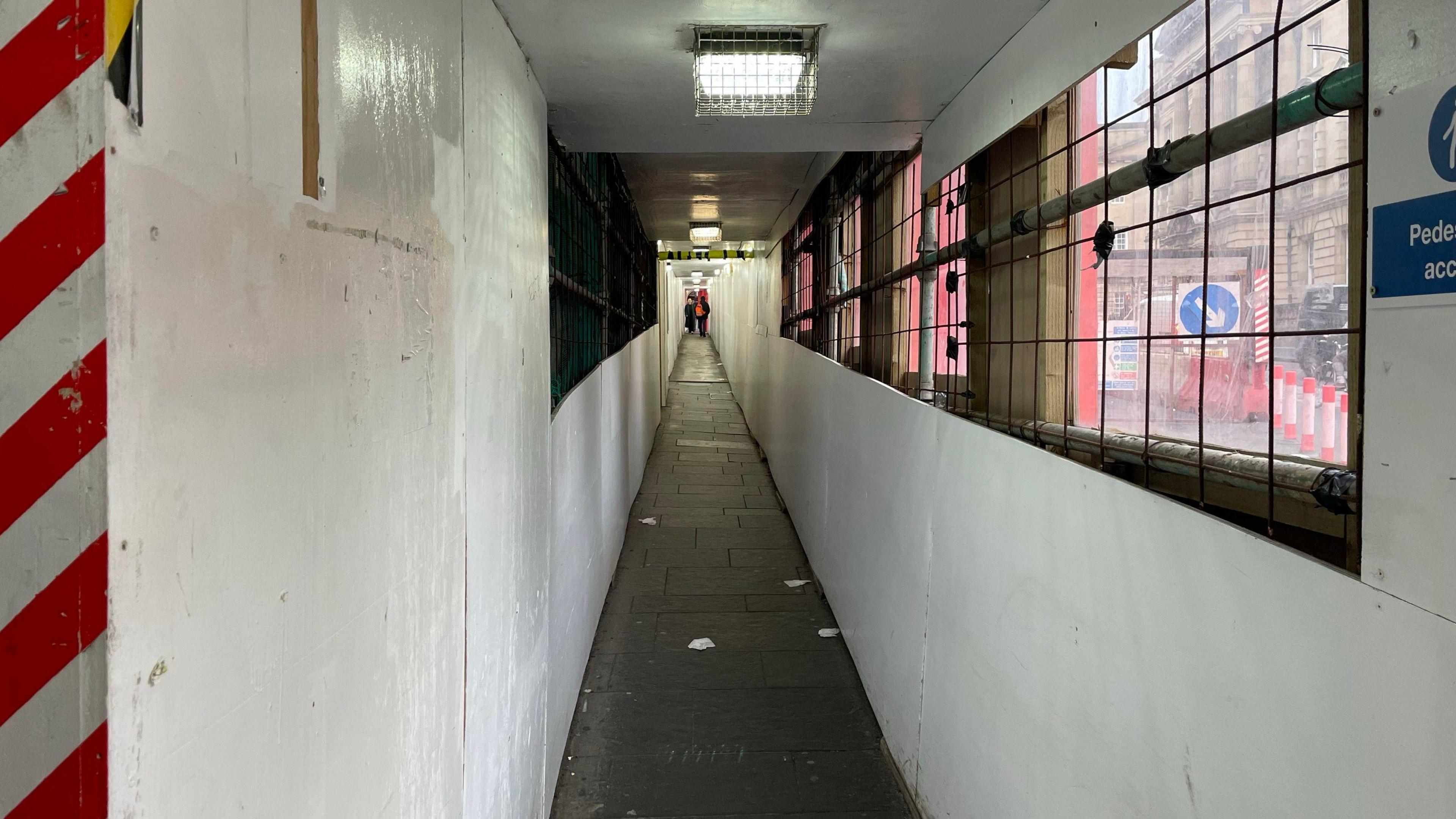 A covered walkway under the G&V Hotel development. The sides are white. Part of the road can be seen through an opening on the right. A ceiling light surrounded by a cage is on the roof.