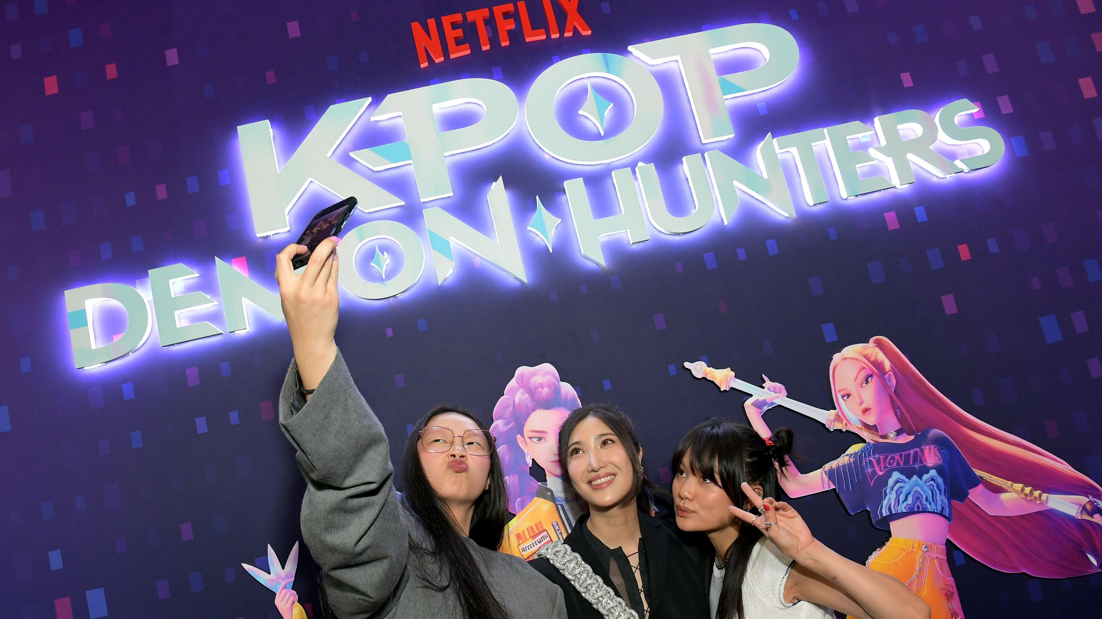 Audrey Nuna, EJAE and Rei Ami attend the KPop Demon Hunters Special Screening at Netflix Tudum Theatre. The woman on the left is holding a camera up in the air for a selfie while the others look up and pose. Behind them is the film's logo and pictures of the animated characters which have long pink and purple hair.