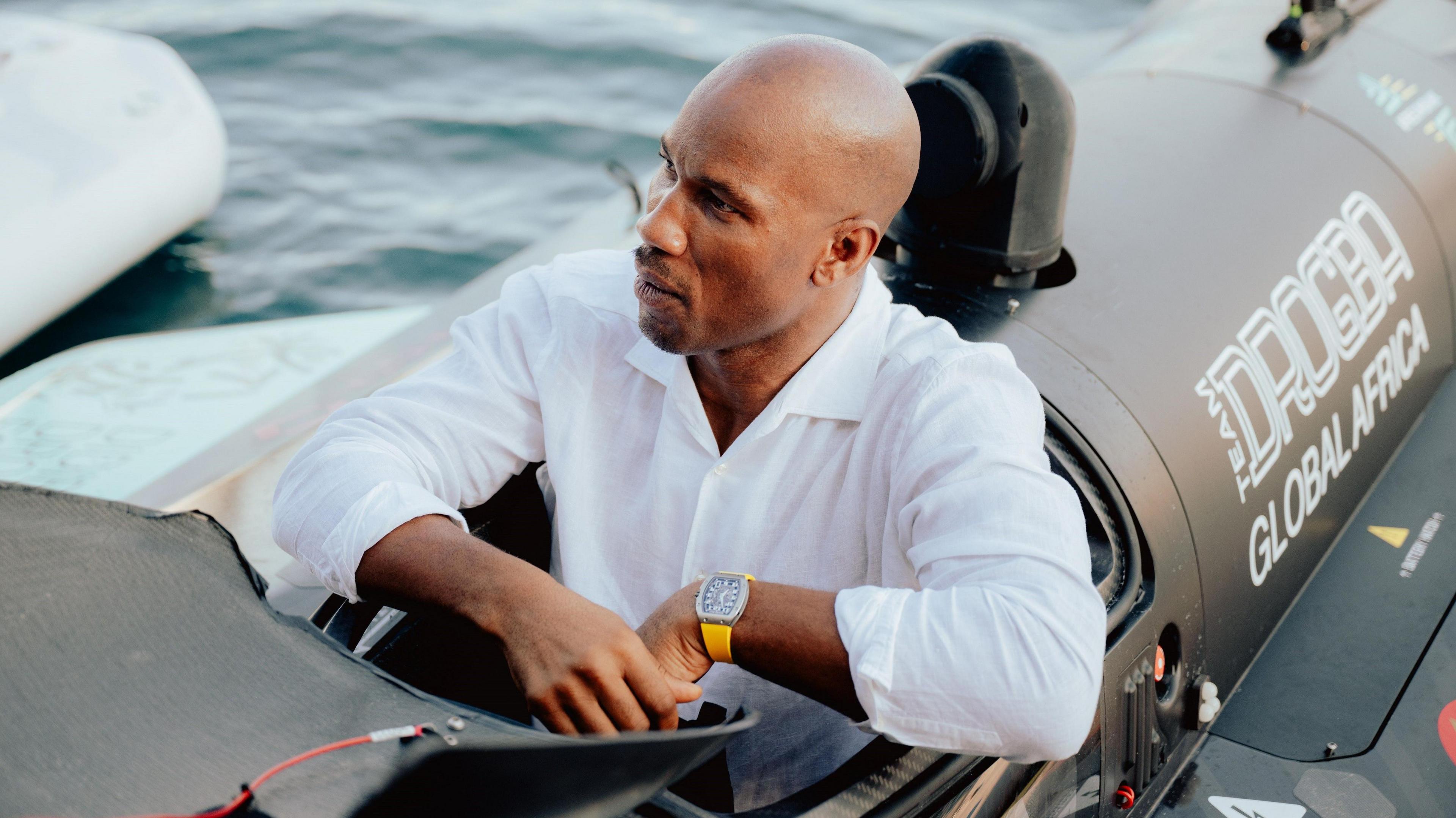 Didier Drogba, wearing a white shirt an watch with bright yellow strap, sits in the cockpit of a black powerboat floating on water which bears the slogan 'Team Drogba Global Africa' on its side