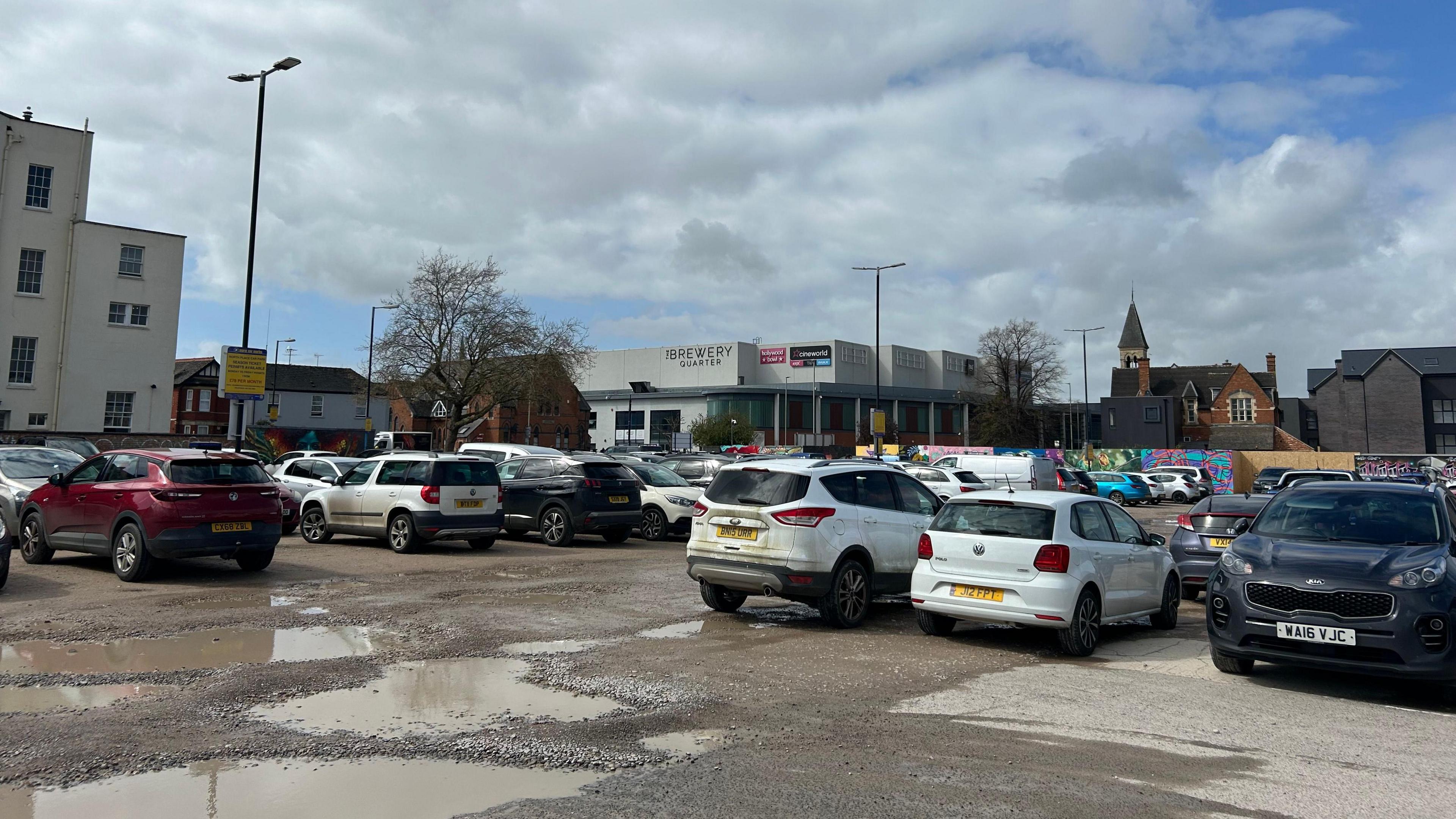 Cars parked in a car park with a wet surface, and water filling the potholes, and a shopping centre in the background