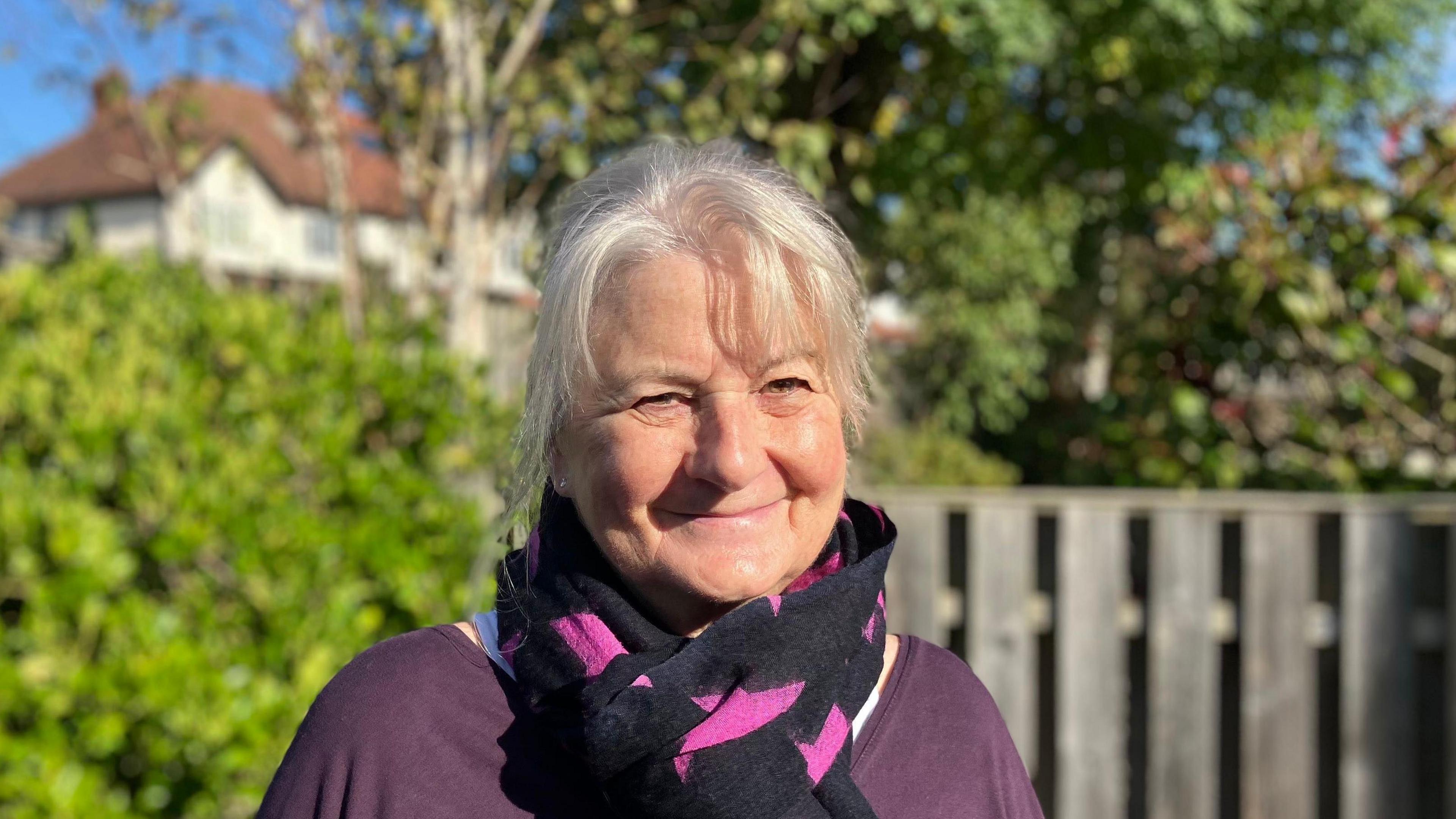 Margaret Simpson, who has grey hair and is wearing a purple top with a purple scarf. She is smiling in the photo.