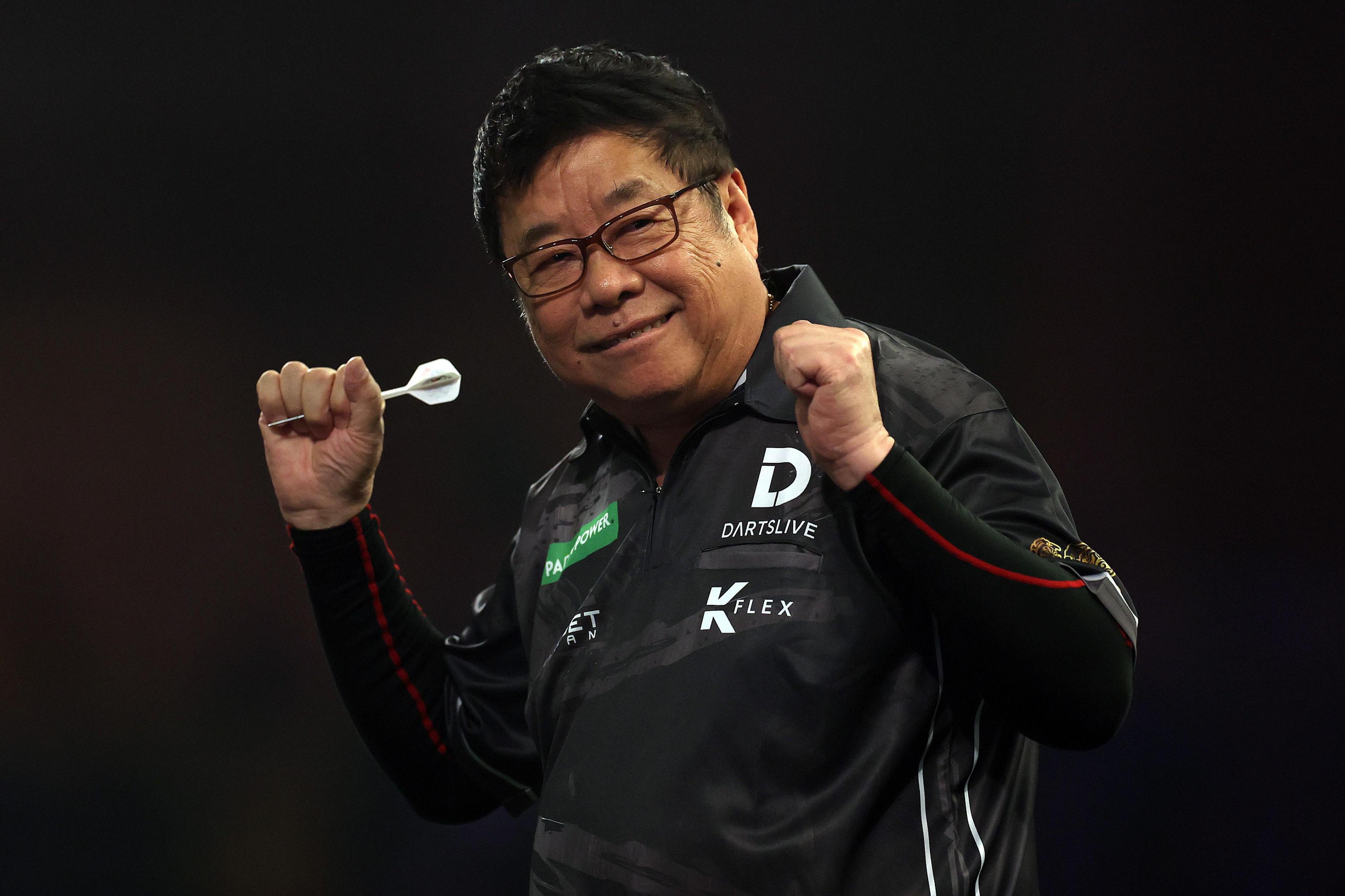 A darts player wearing a black shirt celebrates with clenched fists while holding a dart in one hand, standing under dramatic, low lighting.