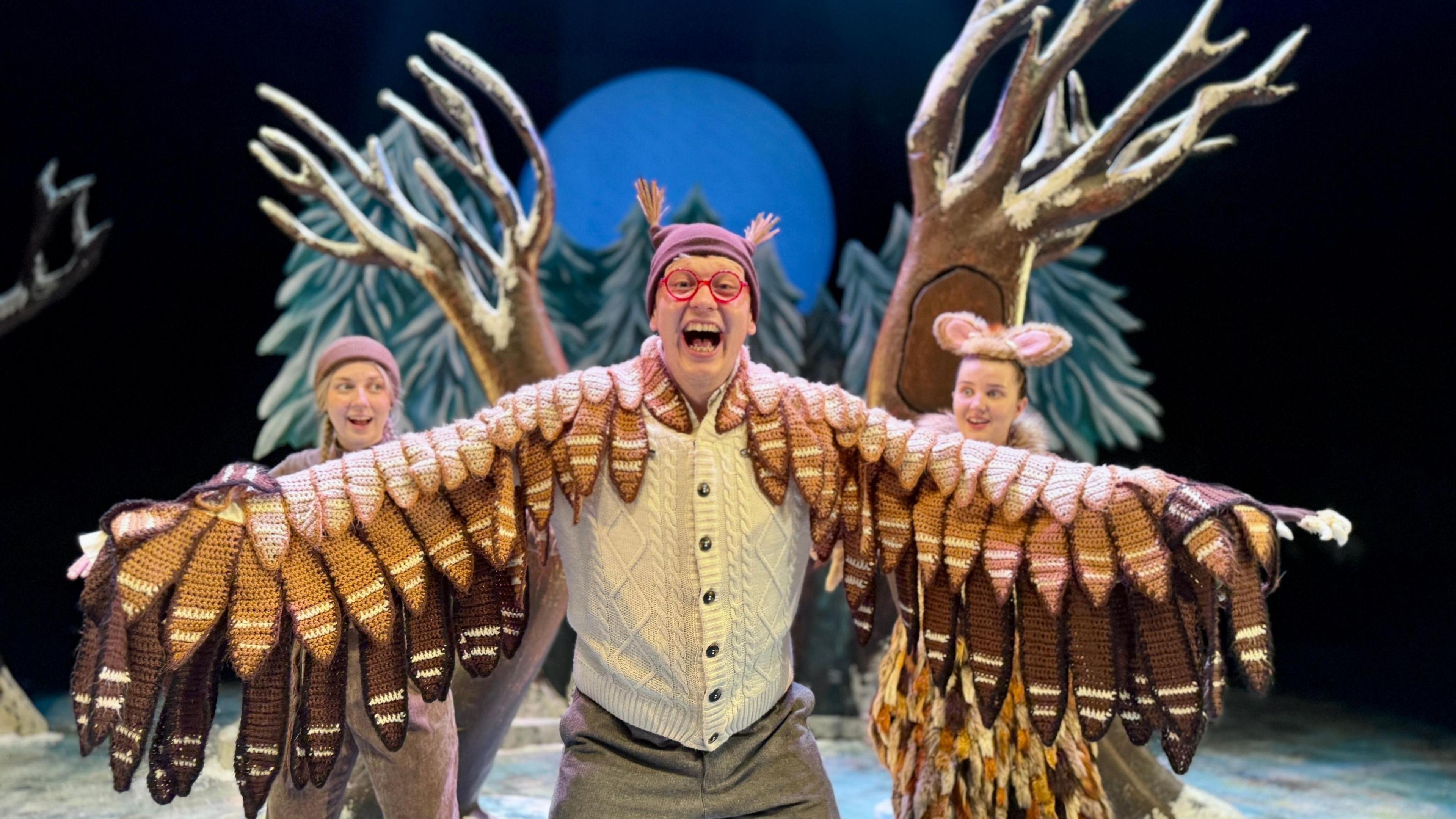 A man in red framed glasses, hat with little tufts for ears, stretches out his arms which have knitted feathers to make it look like he has wings. He is on stage with snowy trees and a moon behind him. Two other characters peer over each wing