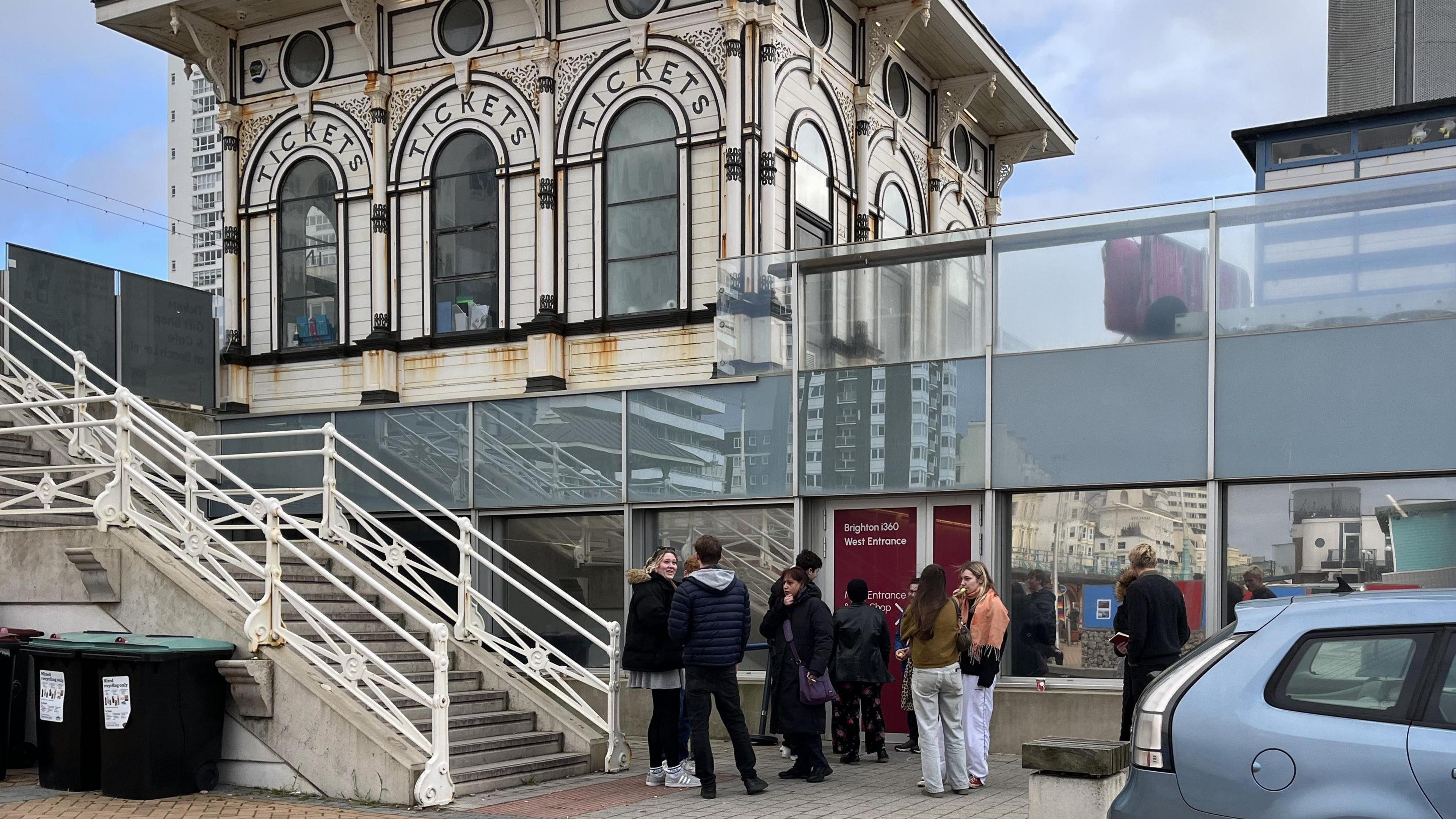 A group of people stand talking to each other outside a building and set of stairs at the bottom of the i360