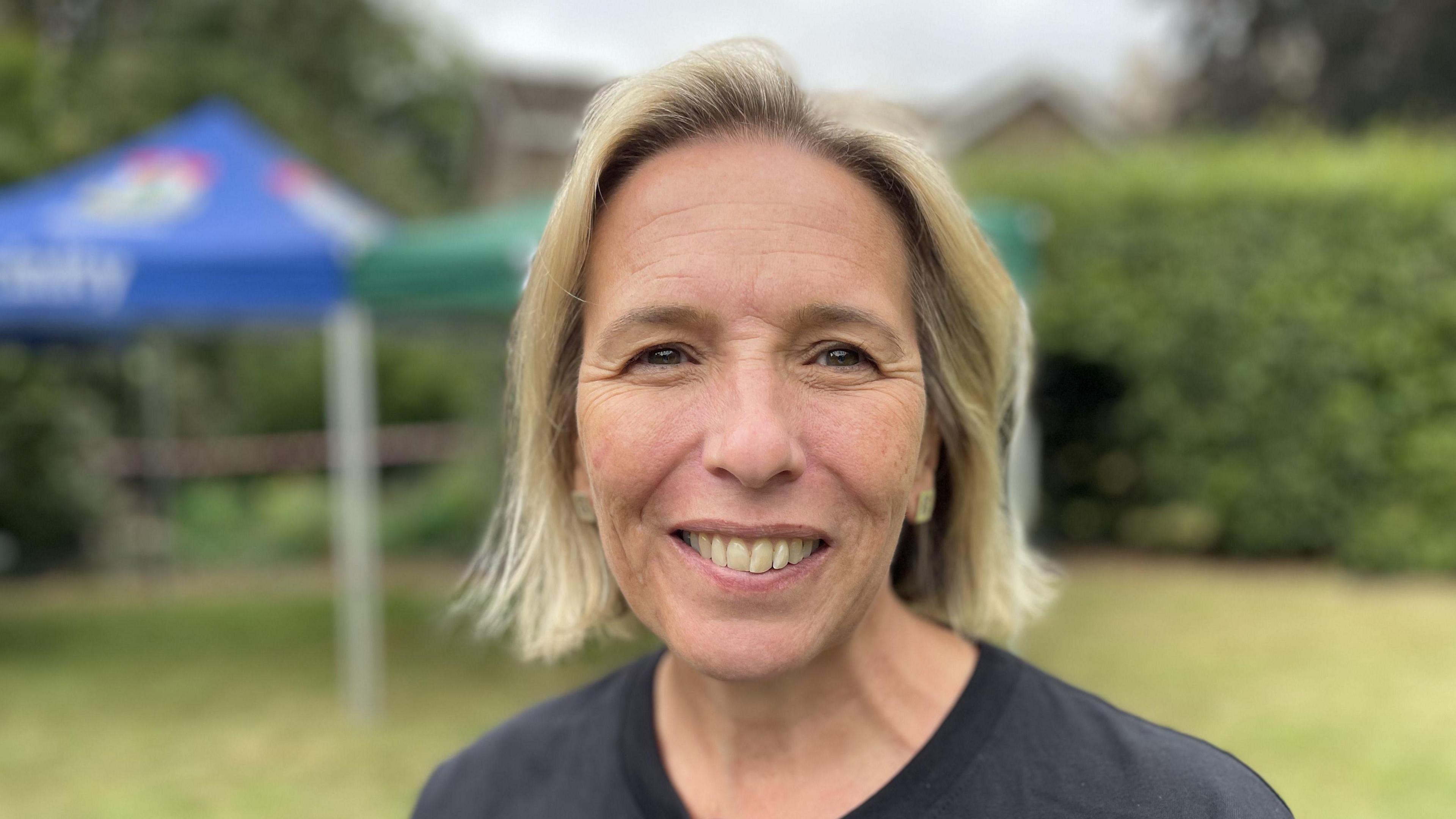 Woman with shoulder length blonde hair and a dark blue top. The background is blurred. She is standing on grass and there is a bush and a marquee behind her.