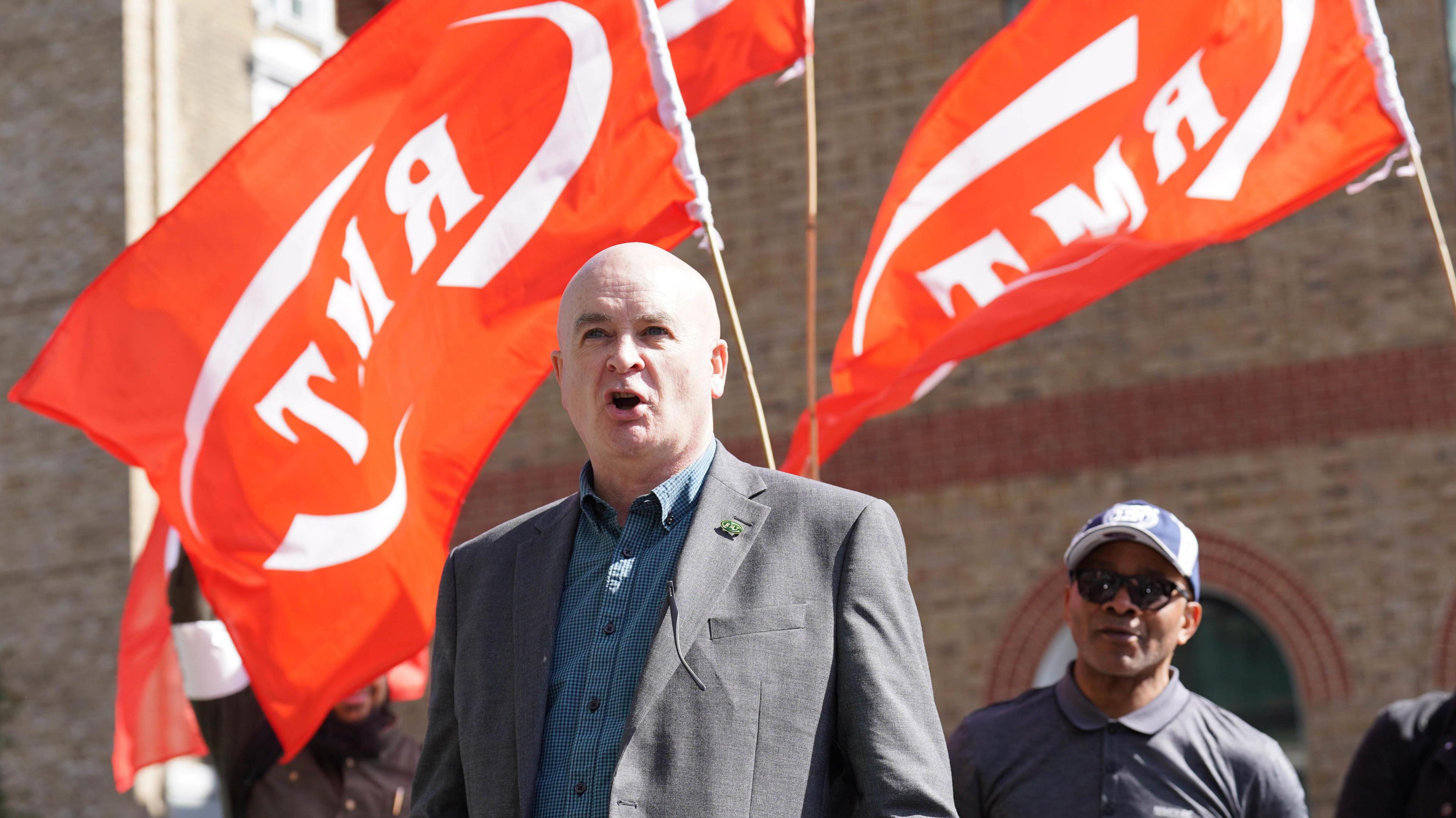 Mick Lynch, who is bald and is wearing a grey suit jacket over a blue shirt, gives a speech as orange RMT flags wave in the backdrop