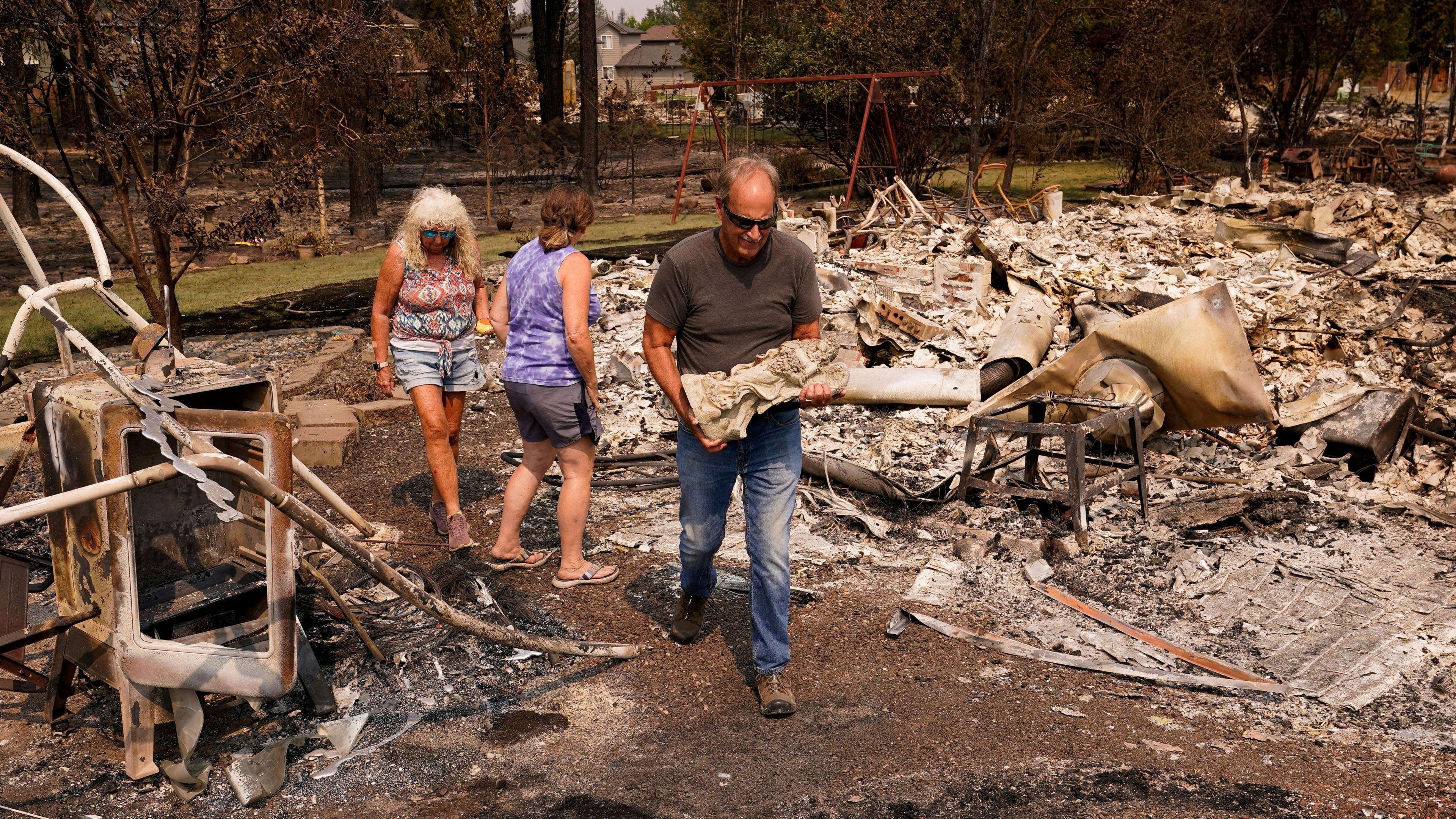 A man carrying a small statue as he walked through burned-out rubble and ashes, with a child's swing seen in a garden in the background - two women wearing vests and shorts are behind him, one turning to look at the ruins 