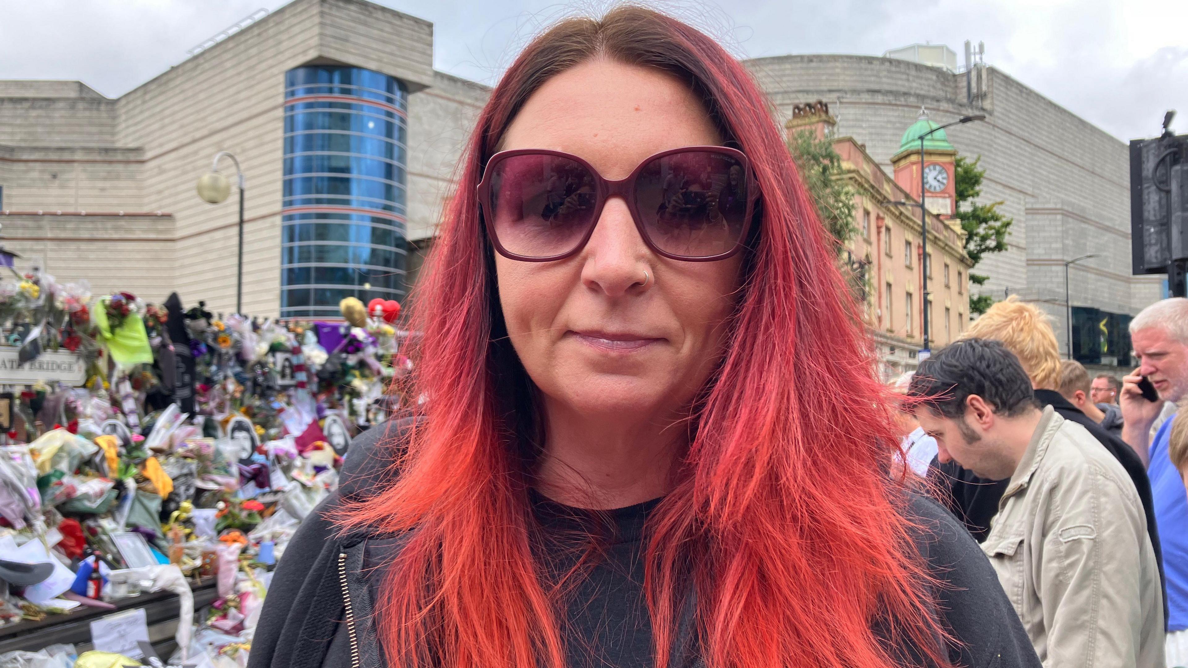 A woman with long red hair, large sunglasses and a black-hooded jumper, stands in front of a pile of floral tributes, while others stand and pay their respects behind her.