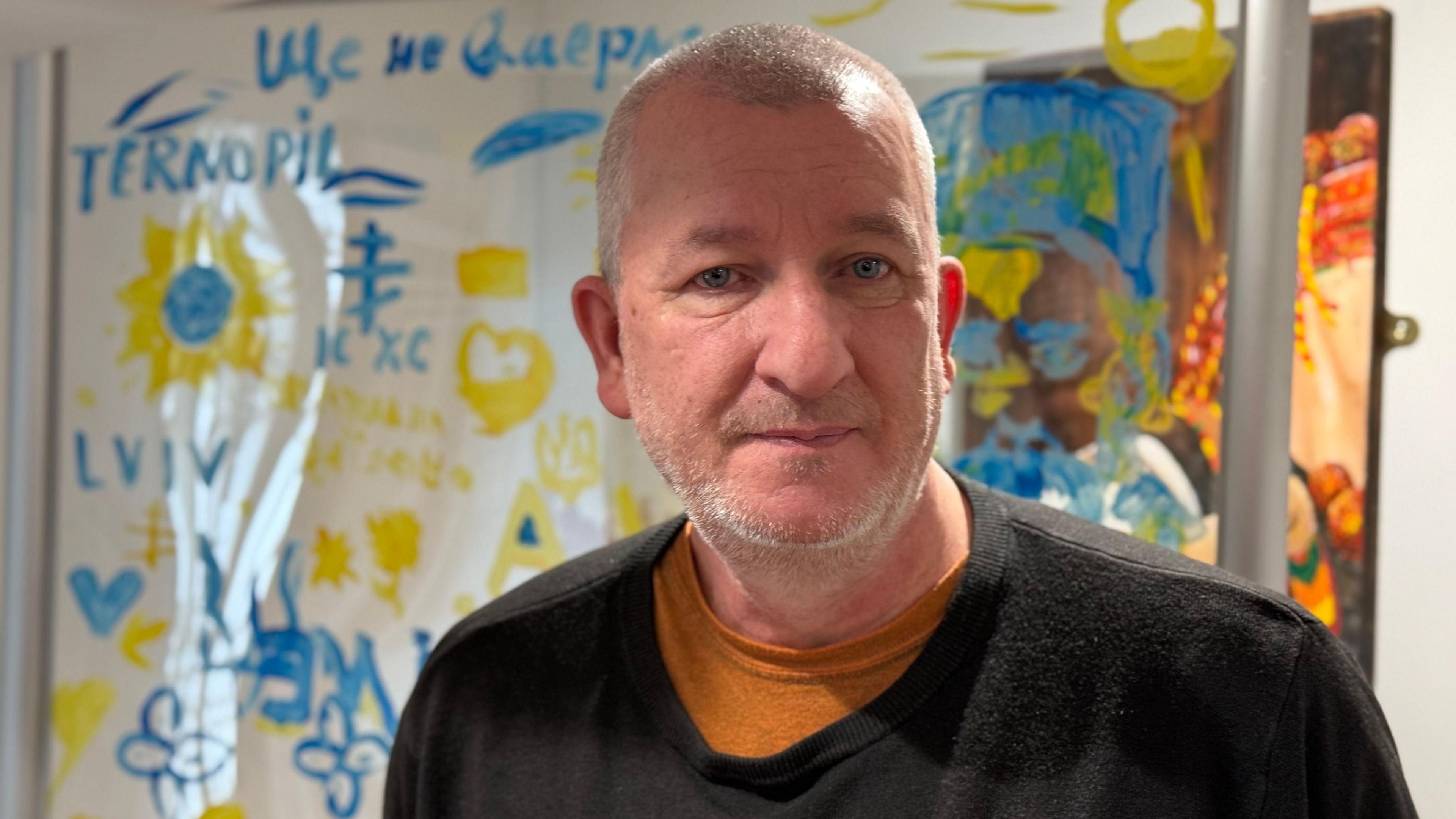 A balding man with grey stubble wearing a black sweatshirt and looking straight at the camera. There's a blue and yellow mural on the wall behind him.