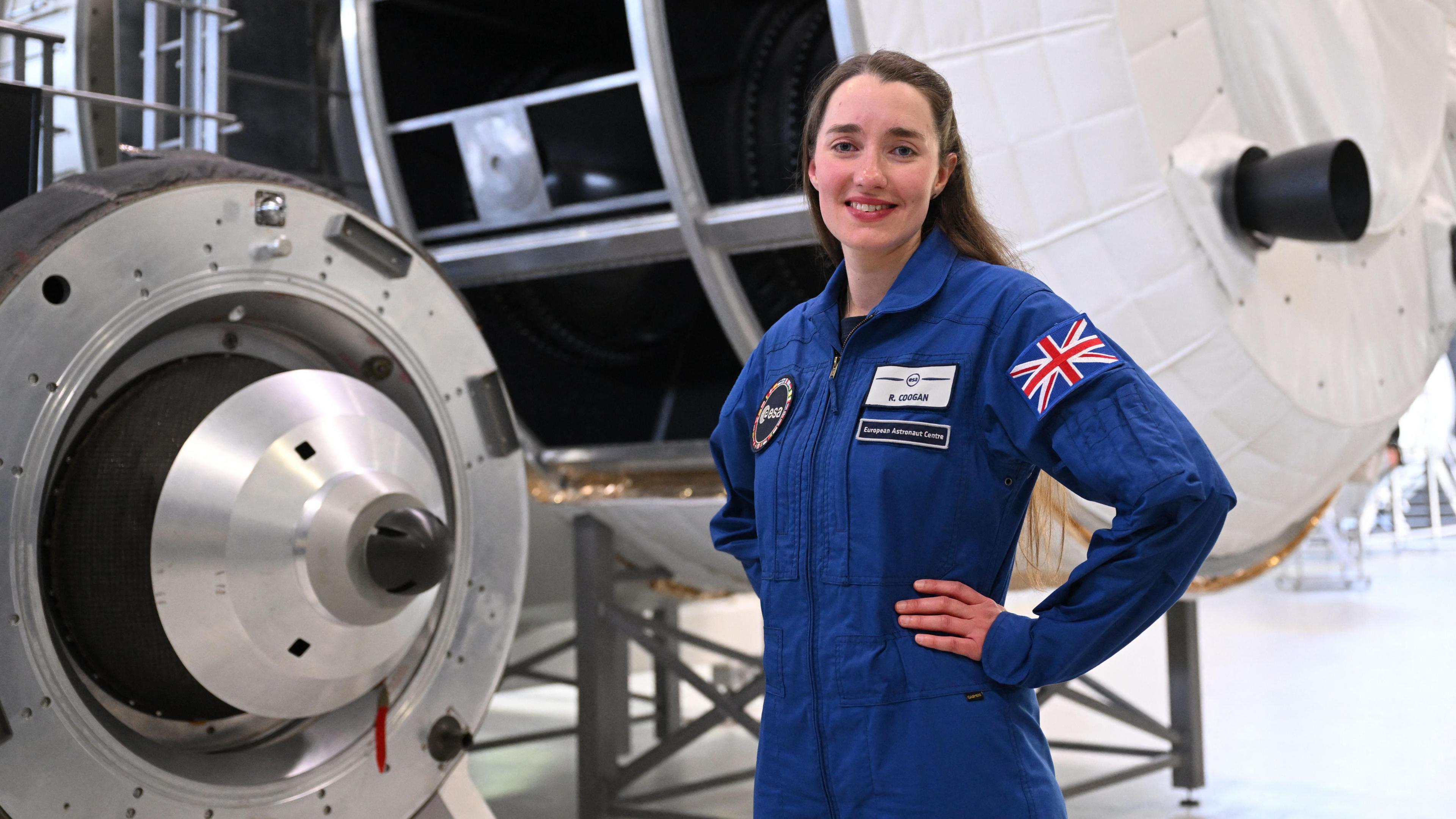 Rosemary Coogan wearing a European Space Agency blue jump suit, has her hands on her hips and is smiling towards the camera. She is stranding in front of two large spacecraft.