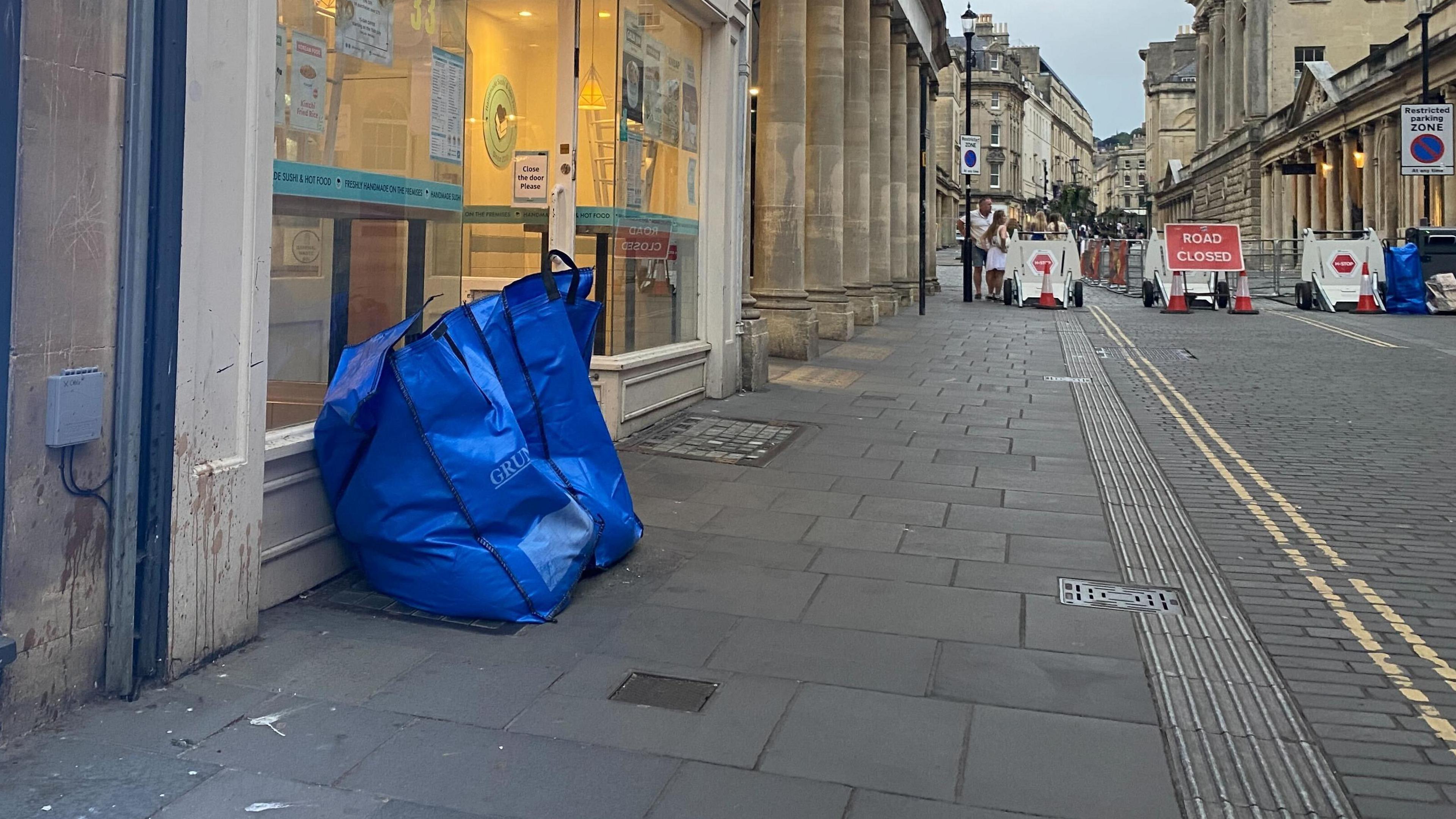 A blue waste bag outside of a business in Bath city centre. In the distance there rea "Road closed" signs and red cones. People can be seen walking in the distance.