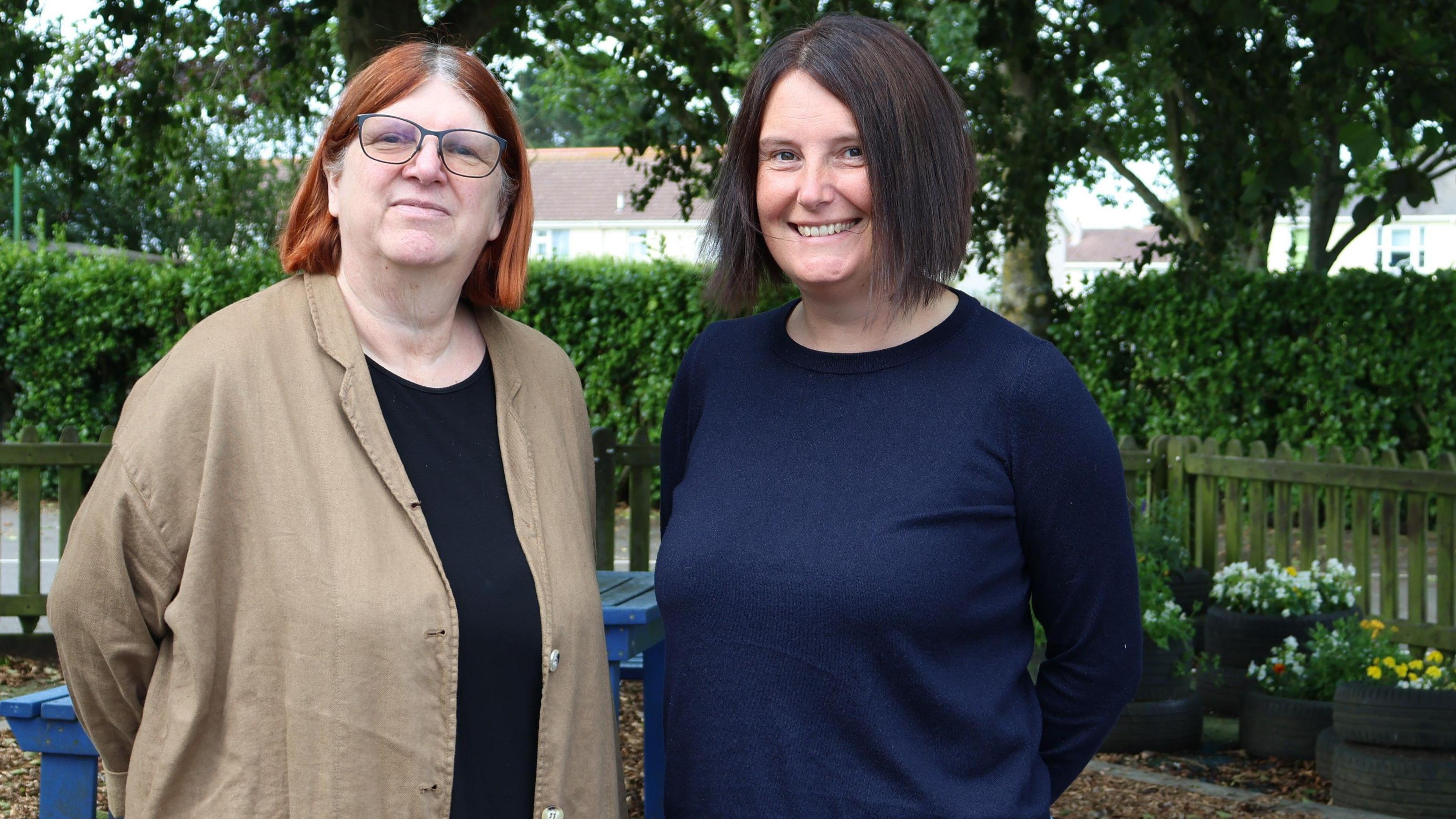 Clare Sealy and Joanne Siddall stand side by side. The pair are standing outdoors. A picket fence, flower pots a green hedge and trees are in the background.