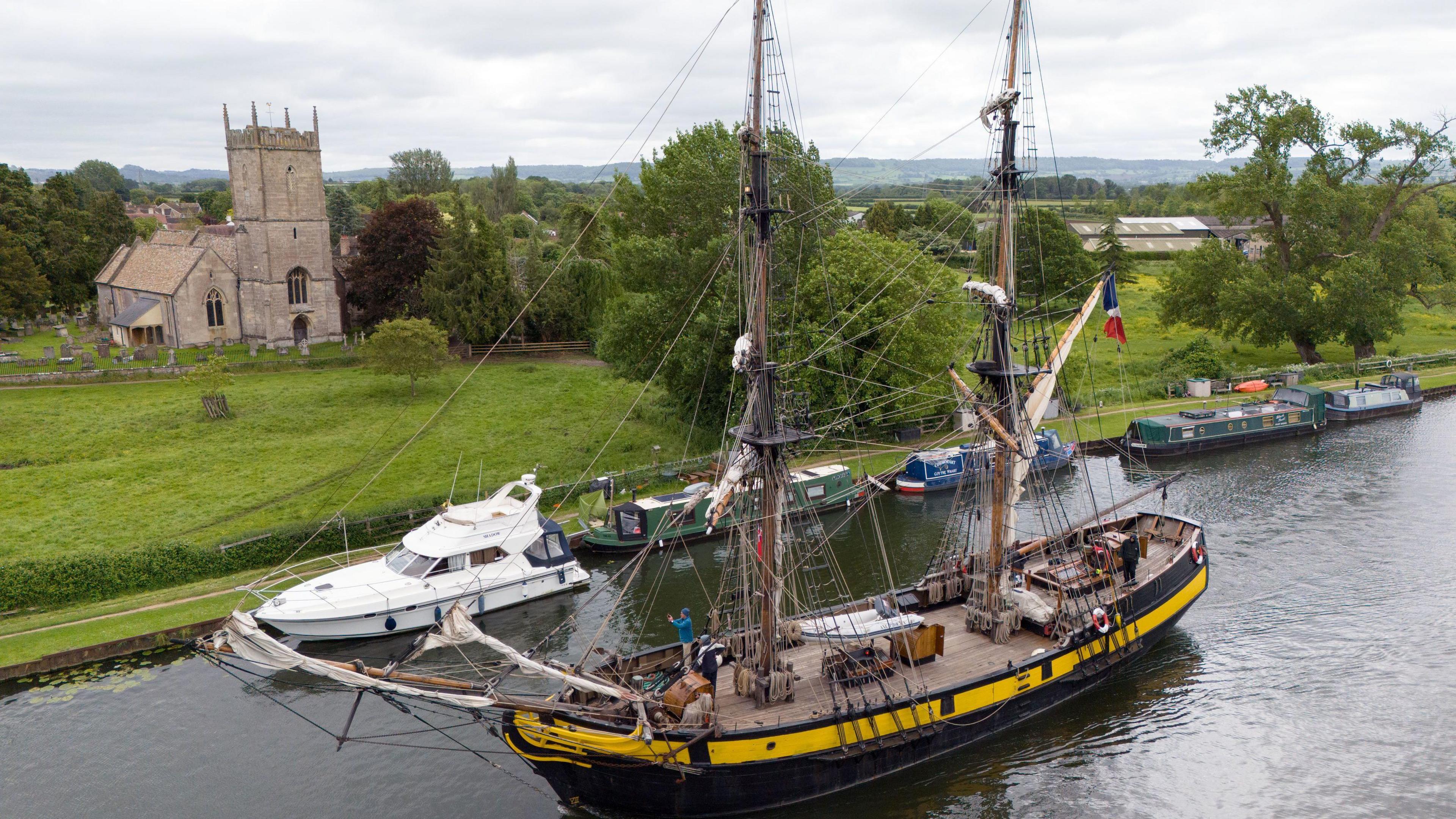 The tall ship Phoenix, which featured in the Ridley Scott film Napoleon, makes it's way along the Gloucester and Sharpness Canal. It has a yellow stripe around it and a flag at the back.