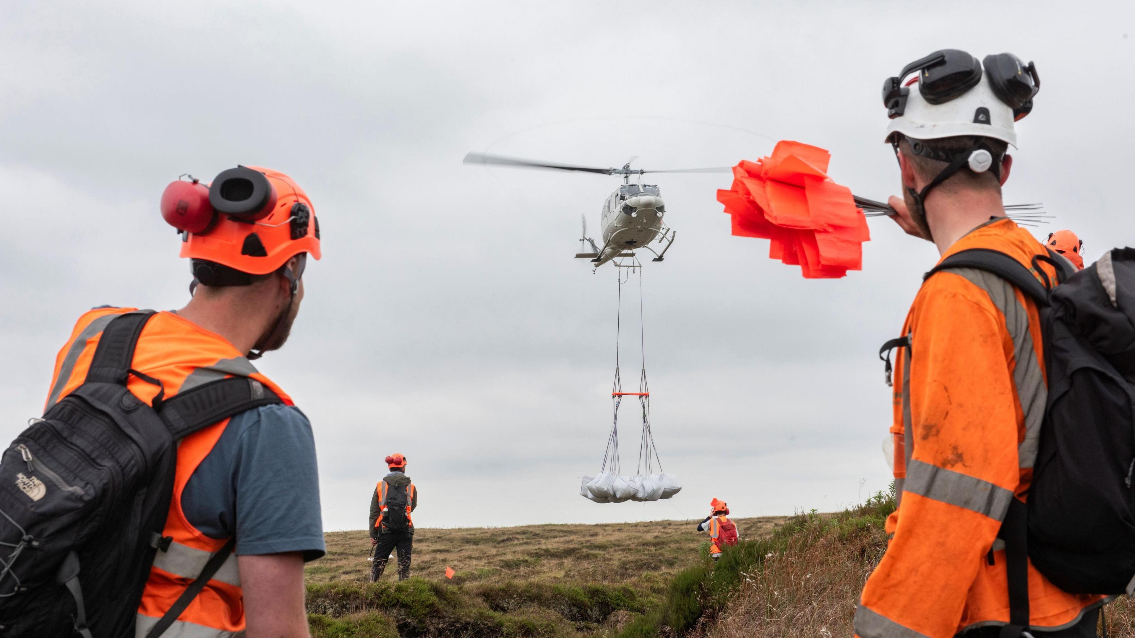 A helicopter carries white bags suspended on a line, marshalled by four people on the ground in hi-vis