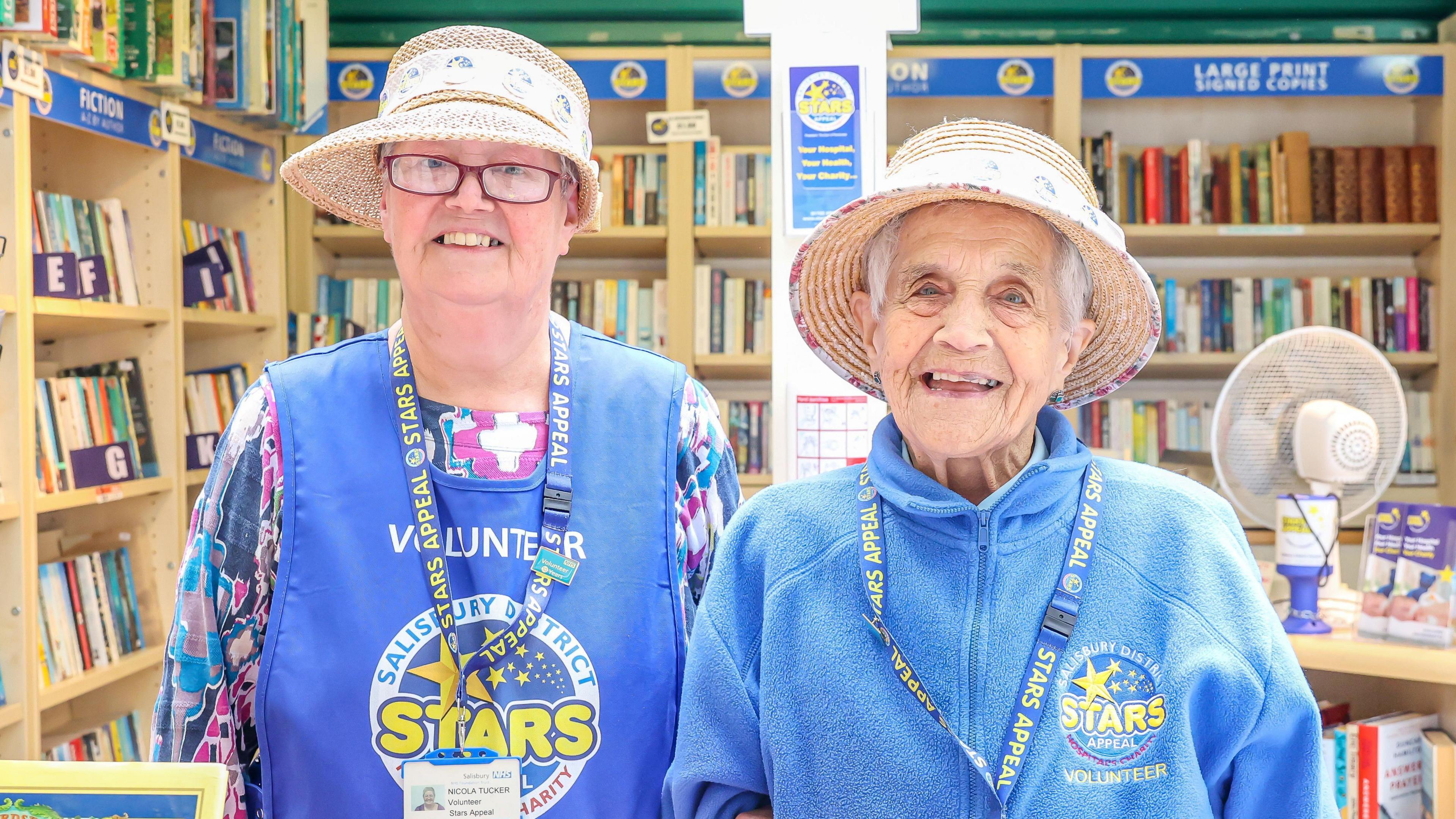 Sybil Tucker and daughter Nicola smile at the camera standing in the Stars Appeal bookshop. They are both wearing straw hats and have a branded clothing and lanyards on