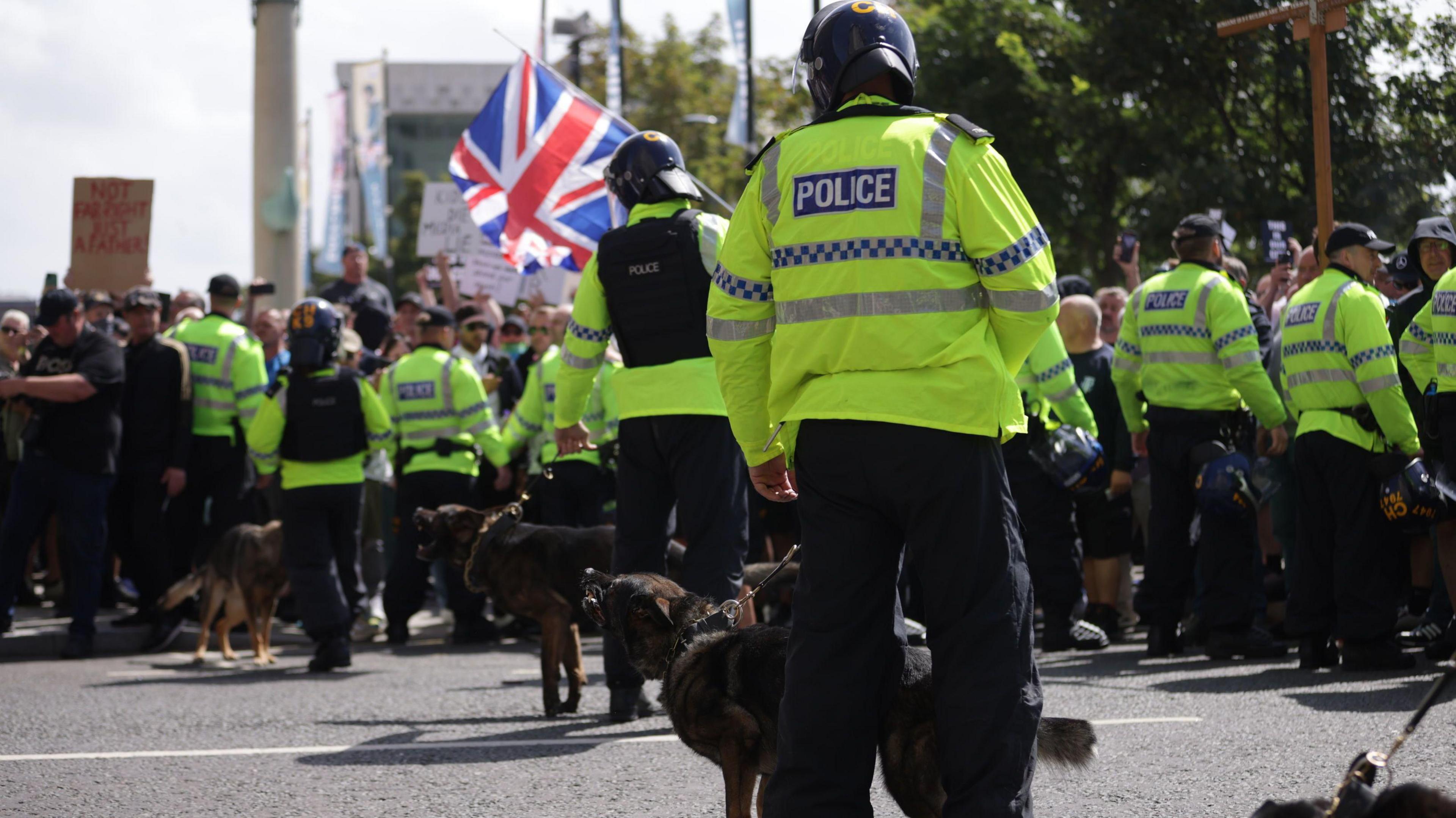 A police dog handler in riot gear and his police dog stand alongside other handlers and dogs and a row of police officers in bright green jackets facing a a group of shouting men and women waving Union Jacks and St George flags