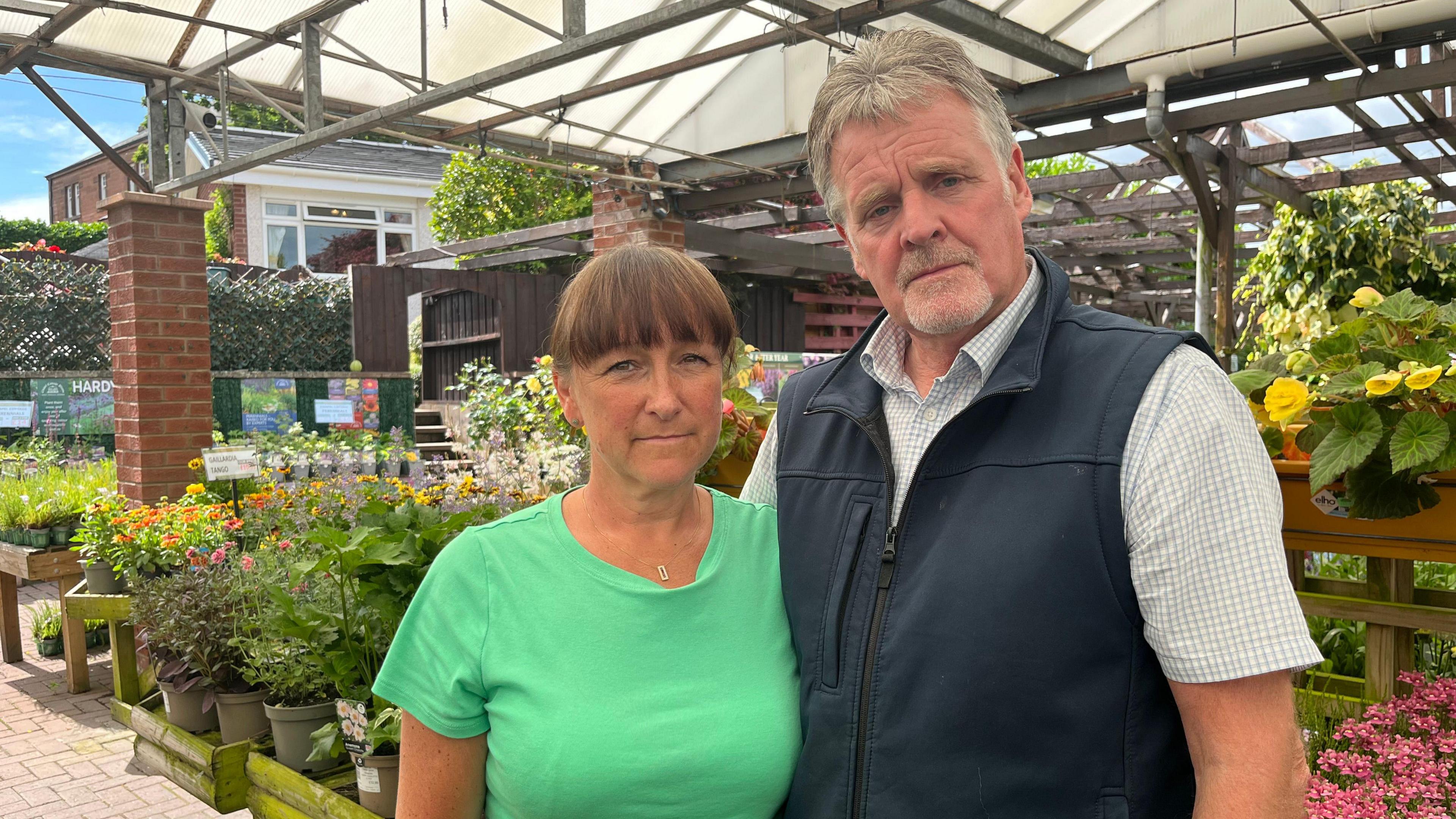 A man and woman look towards the camera - they are stood under a white canopy surrounded by green plants and flowers.