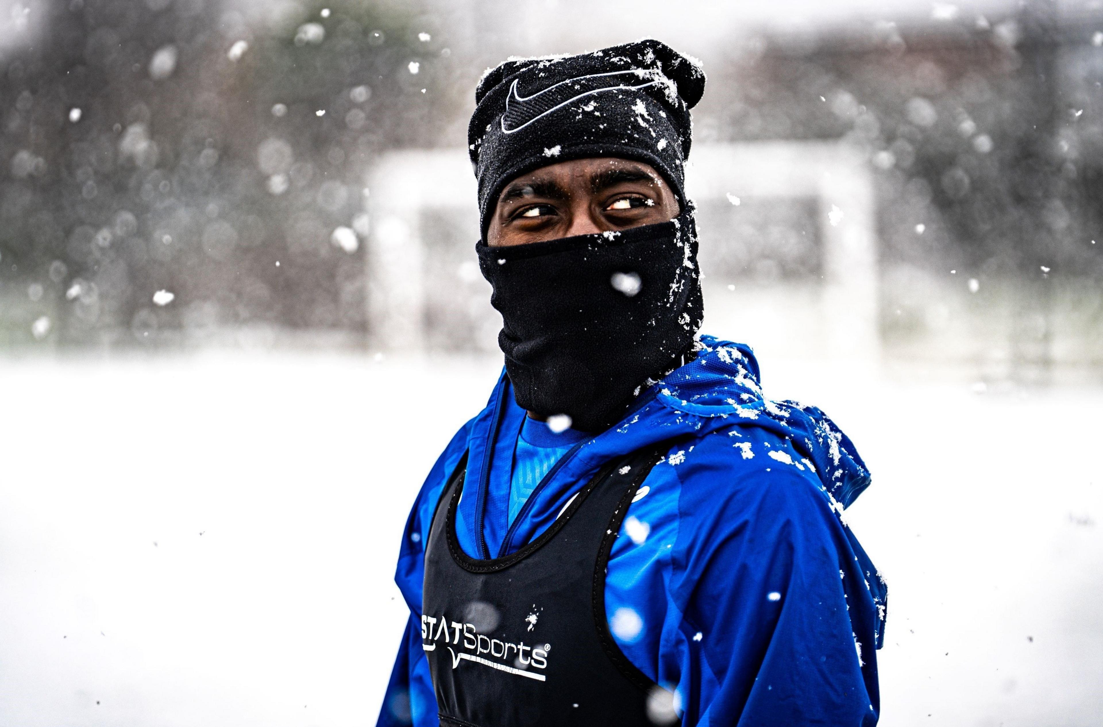 A Bristol Rovers player stands in a training session with heavy snow falling. He has several layers of clothing on, plus a black hat and black snood