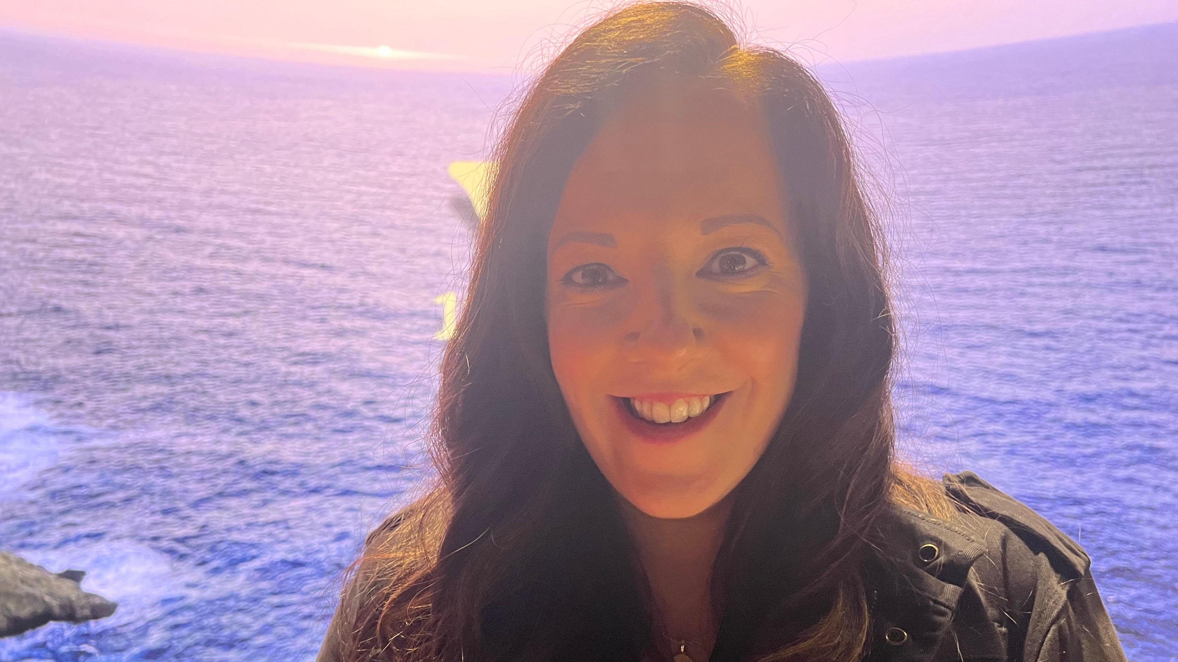 A woman with long brown hair wearing a black jacket smiles at the camera.