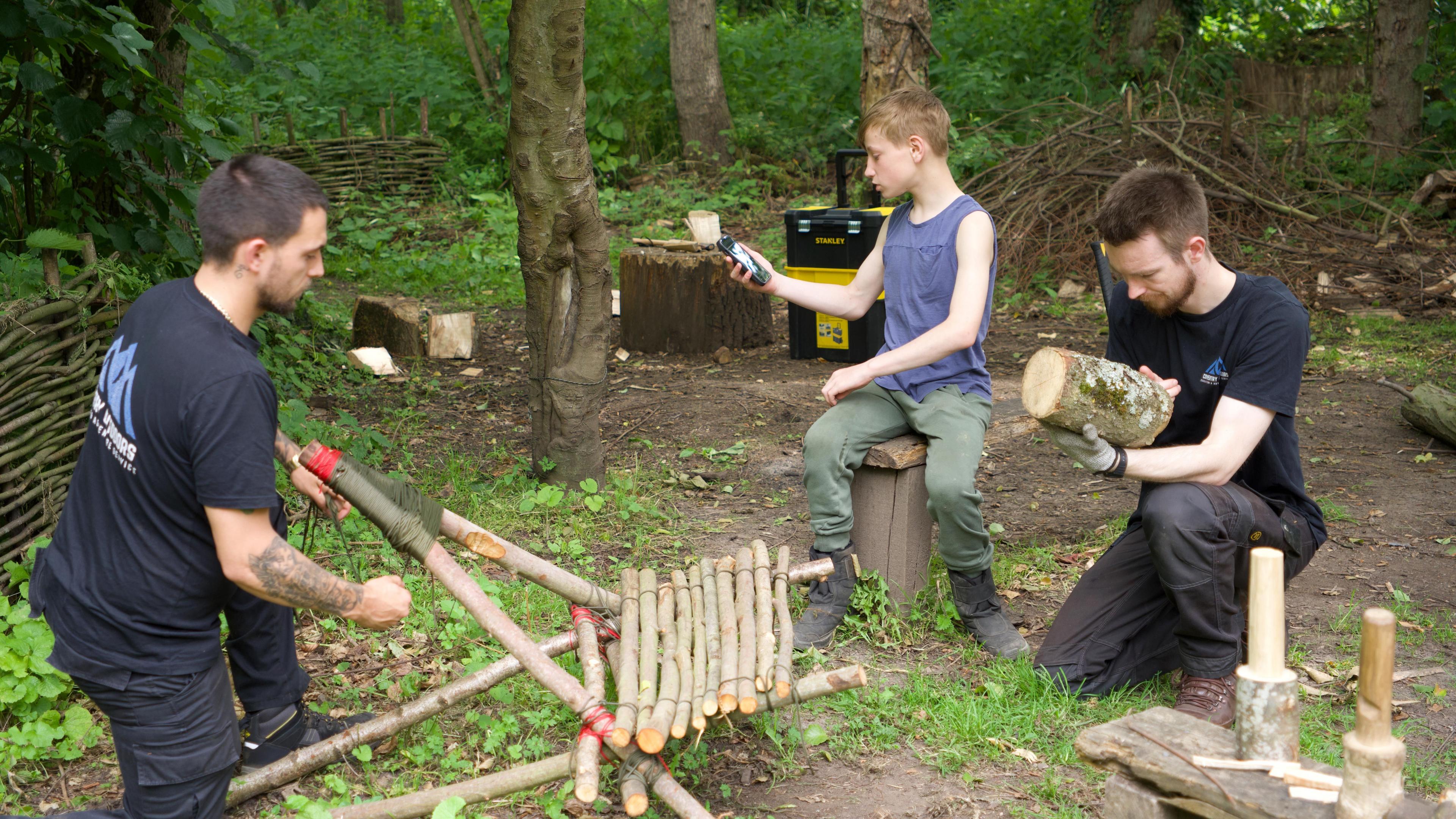 Two men in black t-shirts help a young boy in a blue top and green trousers with woodworking skills. One is making a chair out of branches and another had a block of wood in his hand